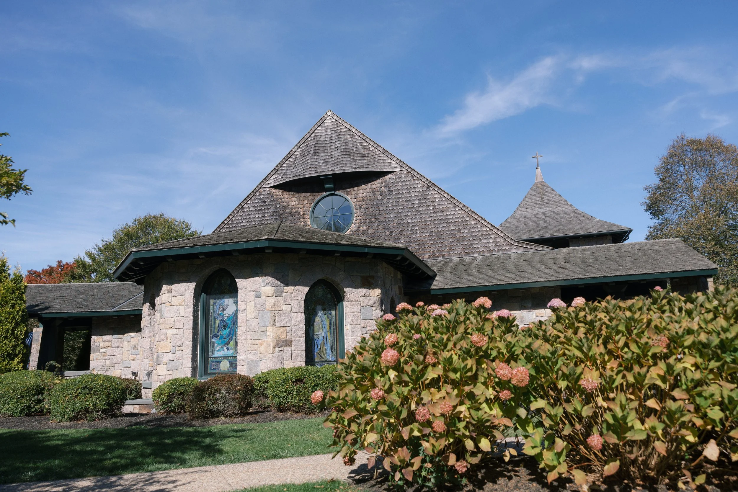 A stone church with stained glass windows, circular window, and a steeple with a cross, surrounded by bushes and trees, under a blue sky.