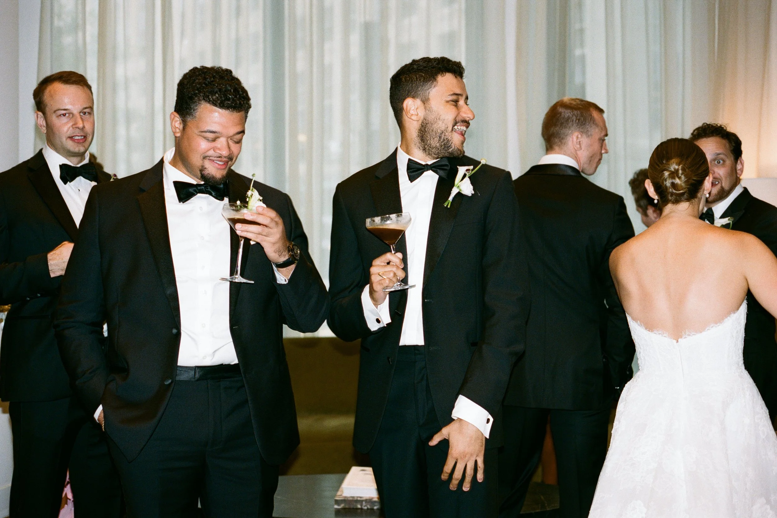 Group of men and women dressed in formal attire at a wedding reception, holding cocktails and chatting.