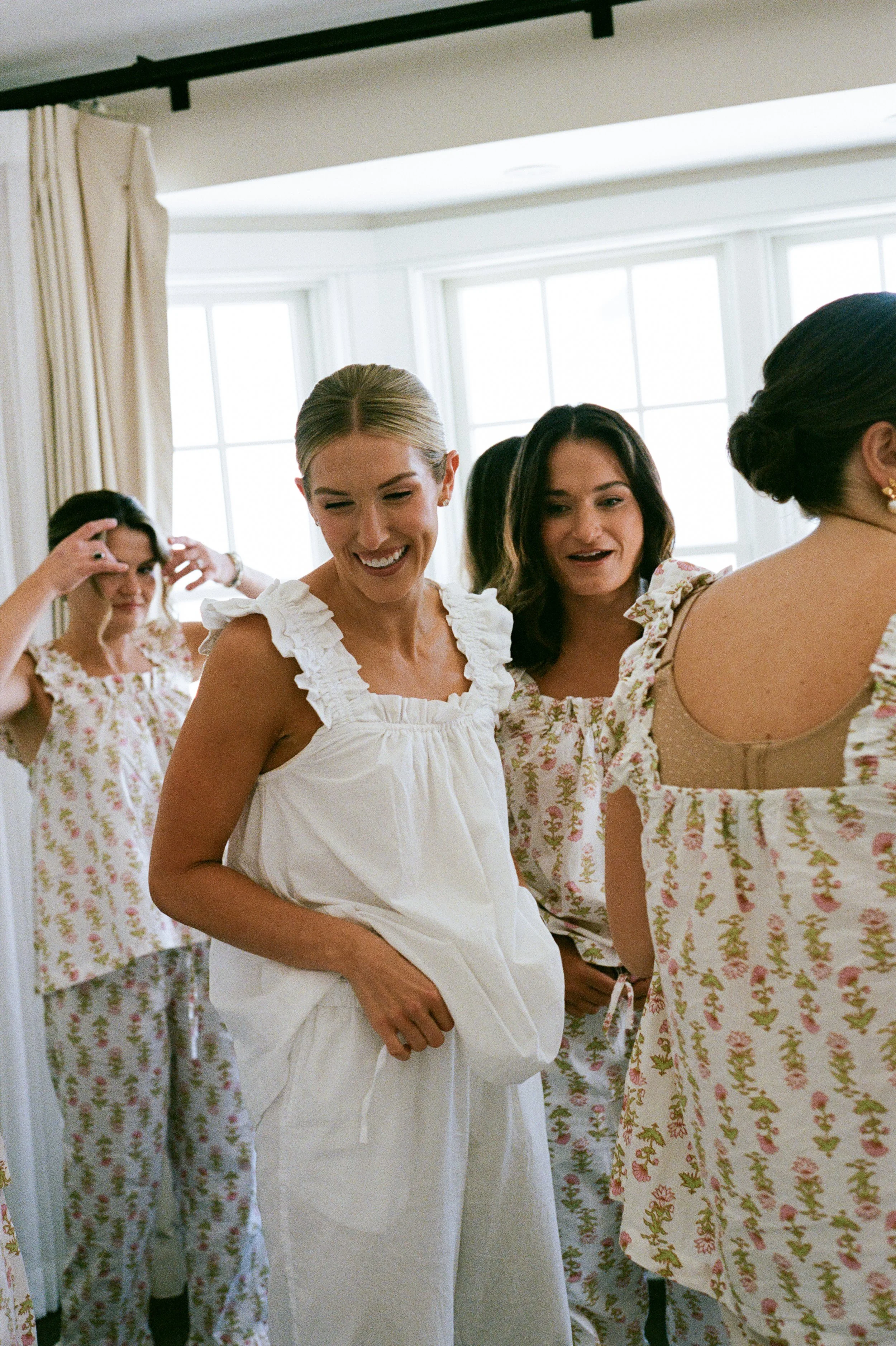 Women in floral pajamas getting ready, smiling, and adjusting their hair, in a room with large windows.