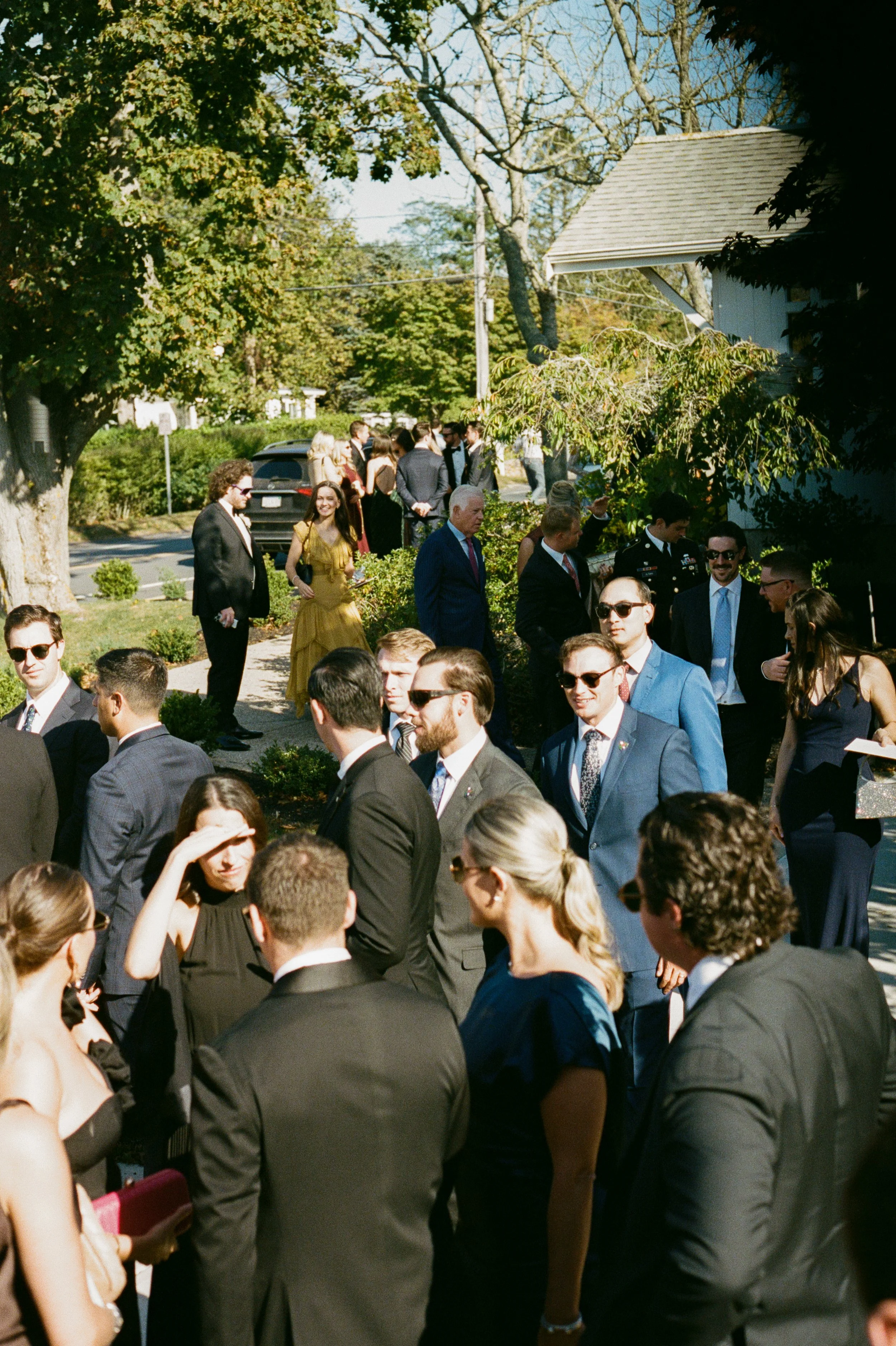 A group of people dressed in formal attire gathered outdoors on a sunny day, likely for a social event or celebration.
