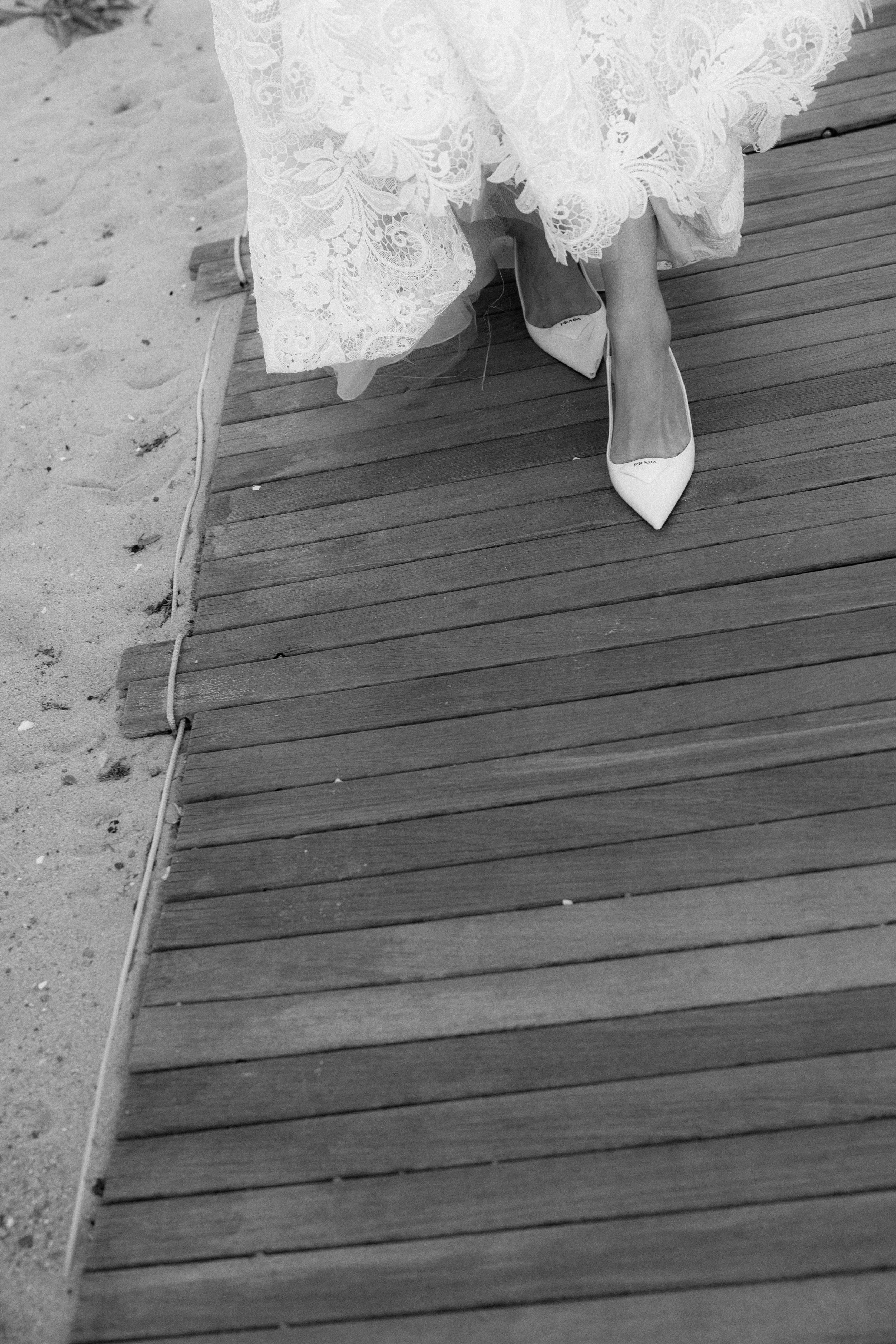 Person wearing white pointed high heels with a lace wedding dress standing on a wooden boardwalk near sandy beach.