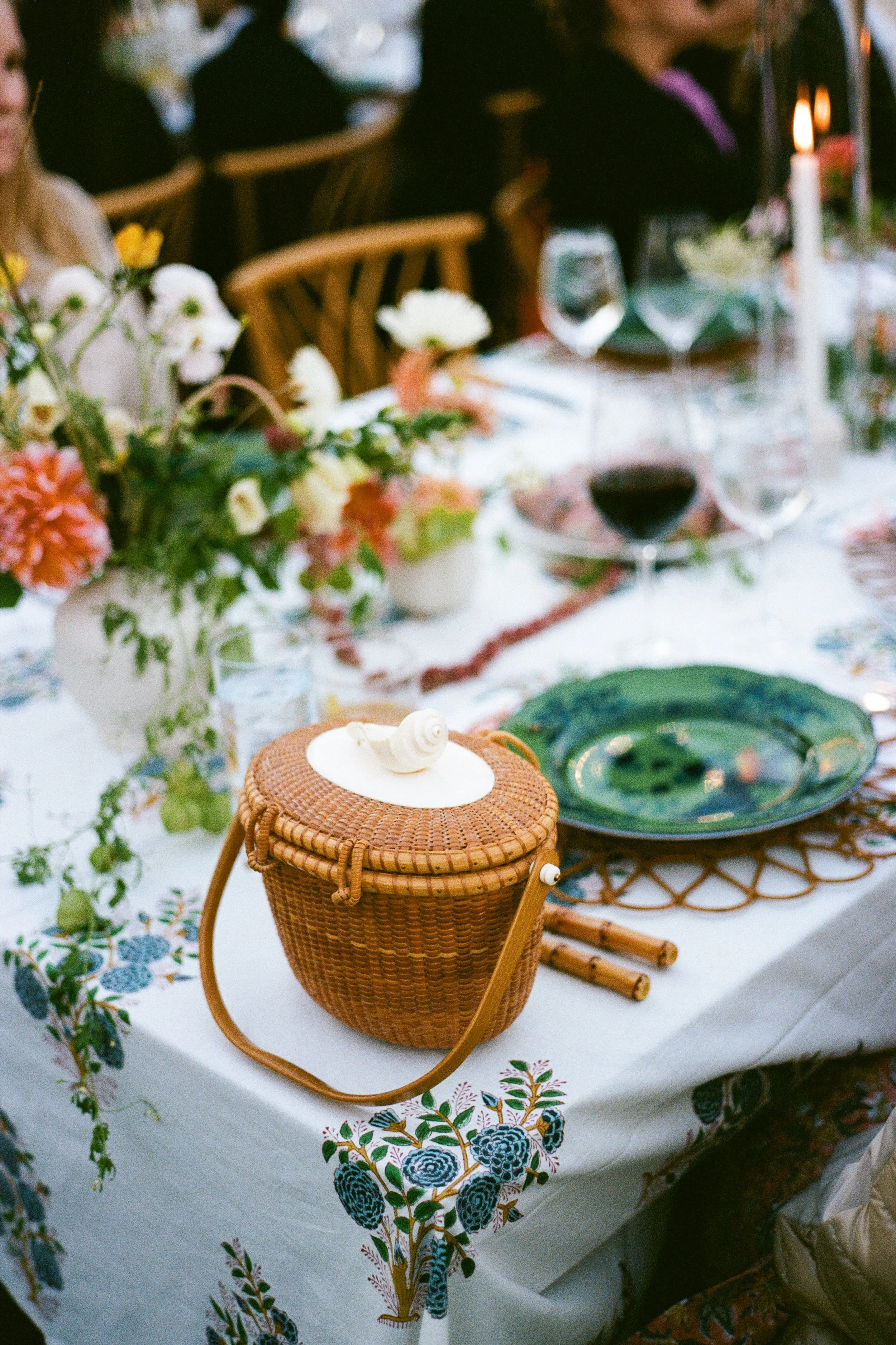 A decorated dining table with floral arrangements, green plates, wine glasses, and a small wicker bag with a snail figurine on top.