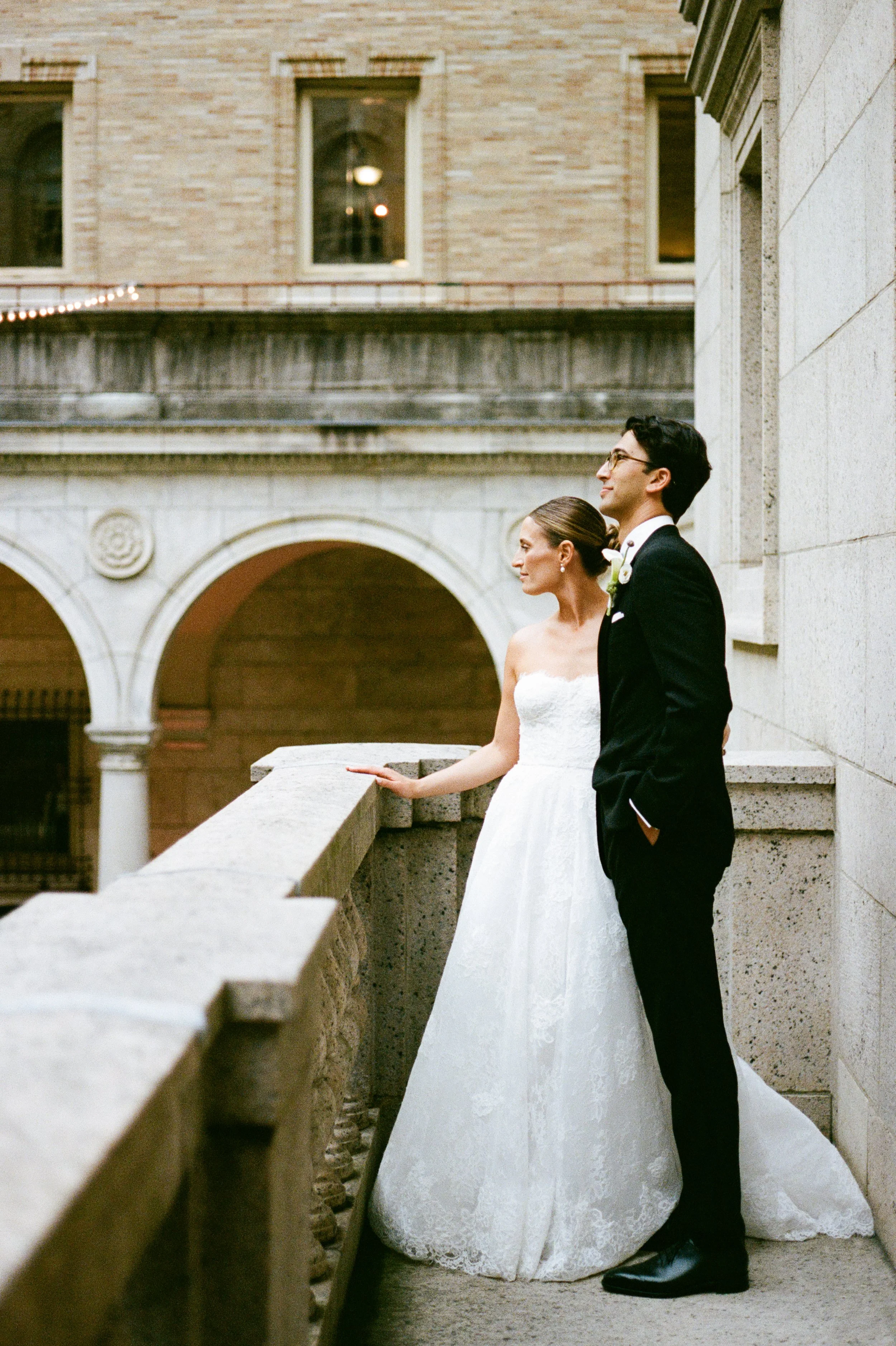 A bride and groom stand on a stone balcony outdoors, dressed in wedding attire, with the bride in a white lace wedding gown and the groom in a black tuxedo, overlooking an architectural backdrop.