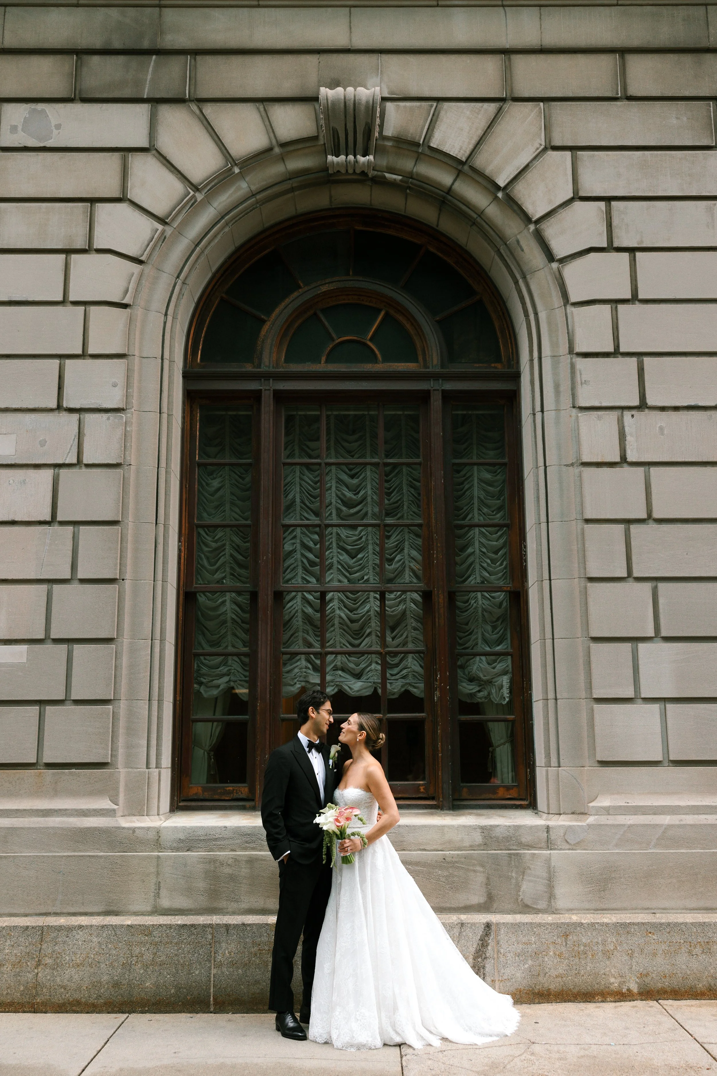 A bride and groom standing close together outside a building with a large arched window. The bride is wearing a white wedding dress and holding a bouquet of pink and white flowers. The groom is dressed in a black tuxedo.