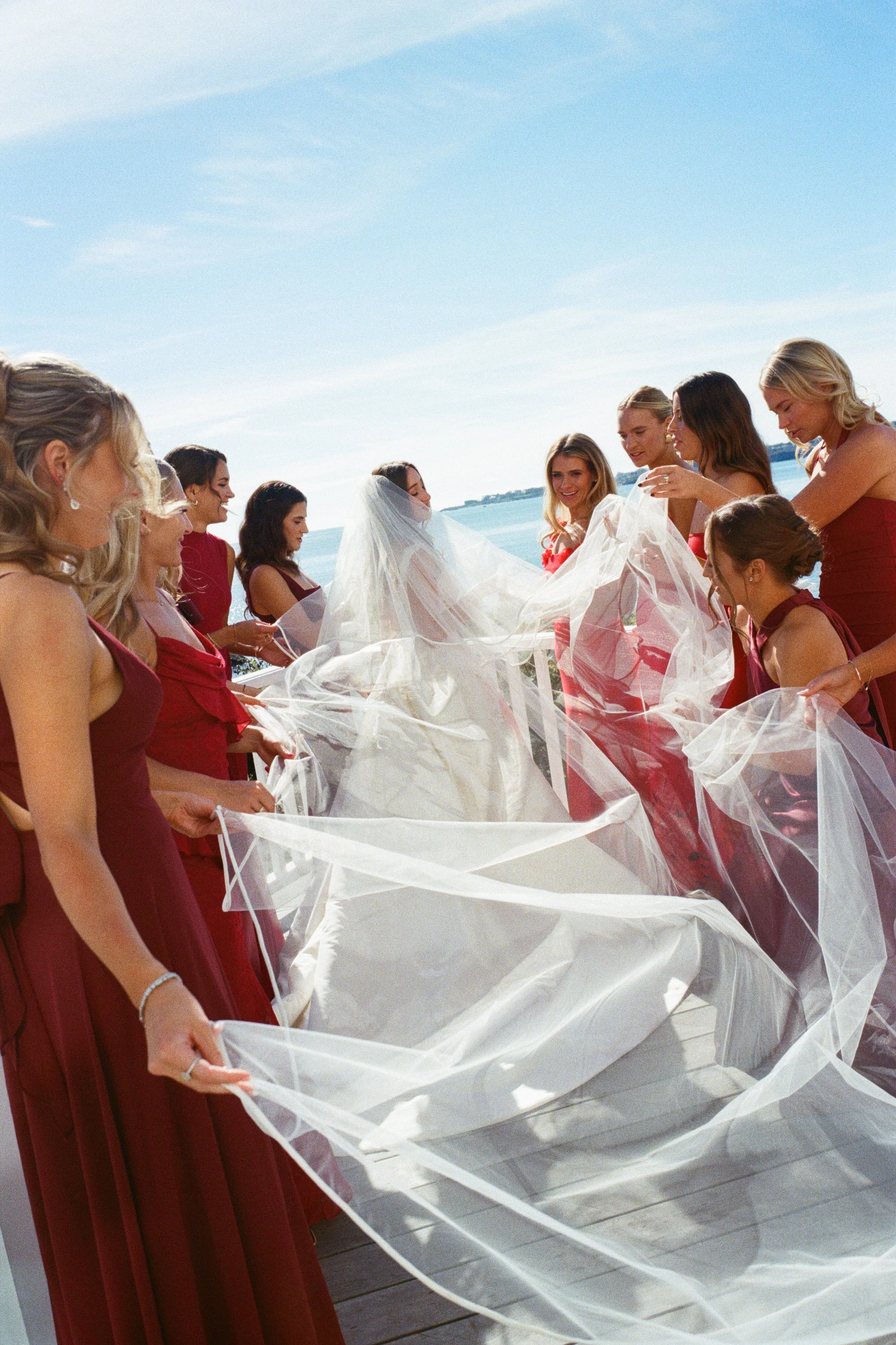 Bride with veil and wedding dress surrounded by bridesmaids in red dresses on a waterfront dock.