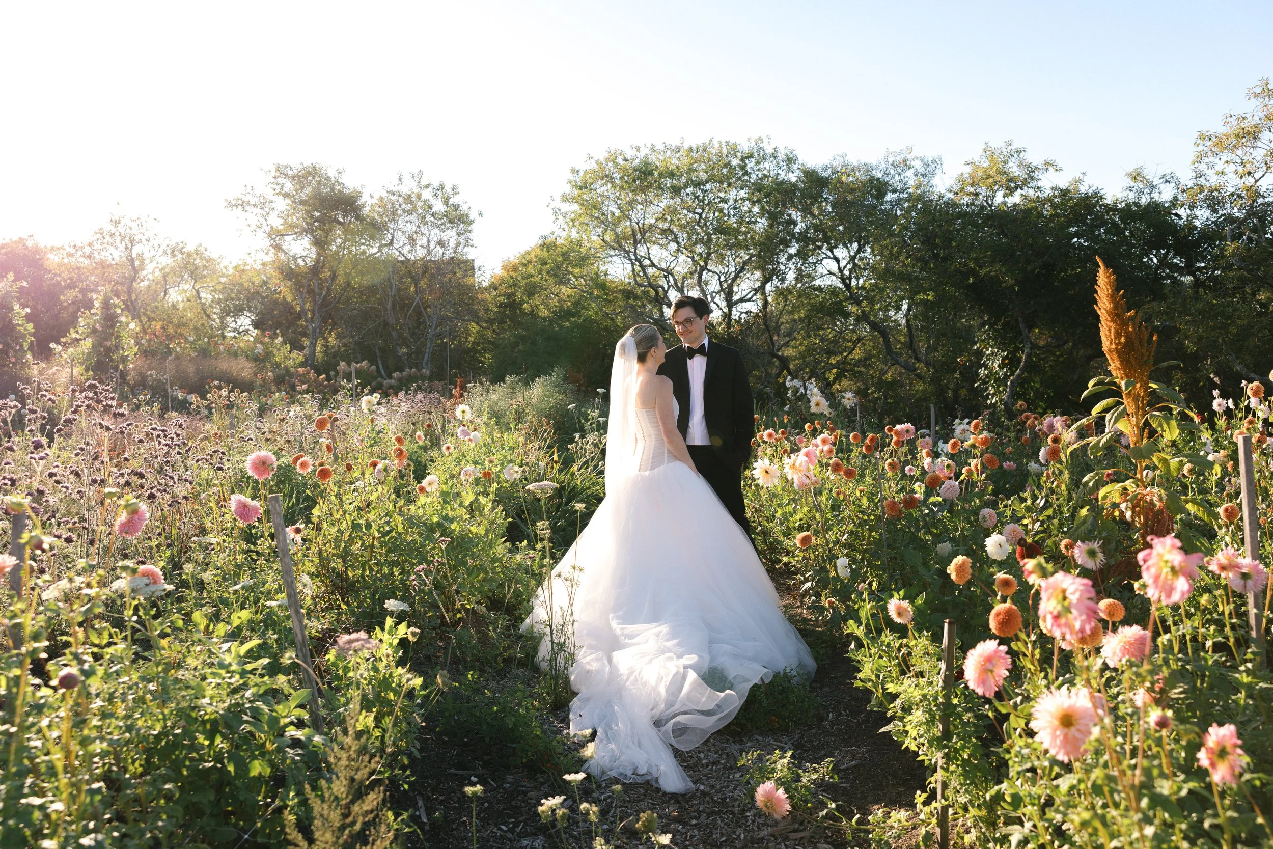 A bride and groom standing in a flower field, facing each other and smiling, with trees and sunlight in the background.
