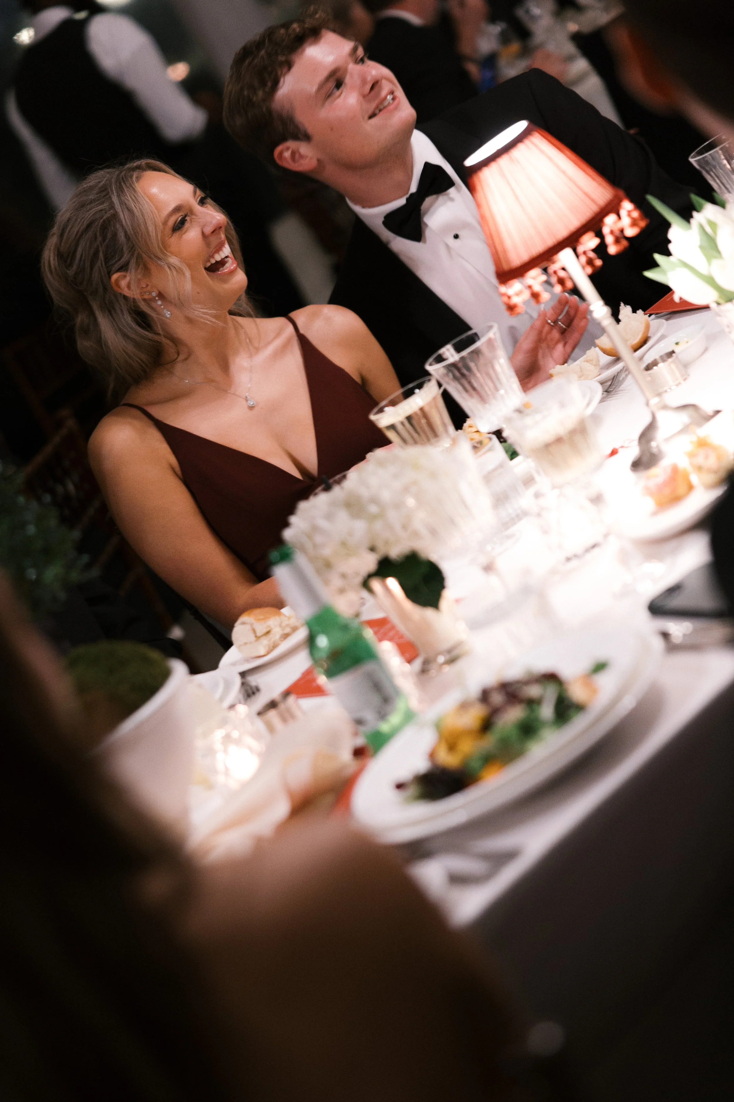 A man in a tuxedo and a woman in a burgundy dress laughing and enjoying a conversation at a formal dinner table decorated with flowers, candles, and food during an event or celebration.