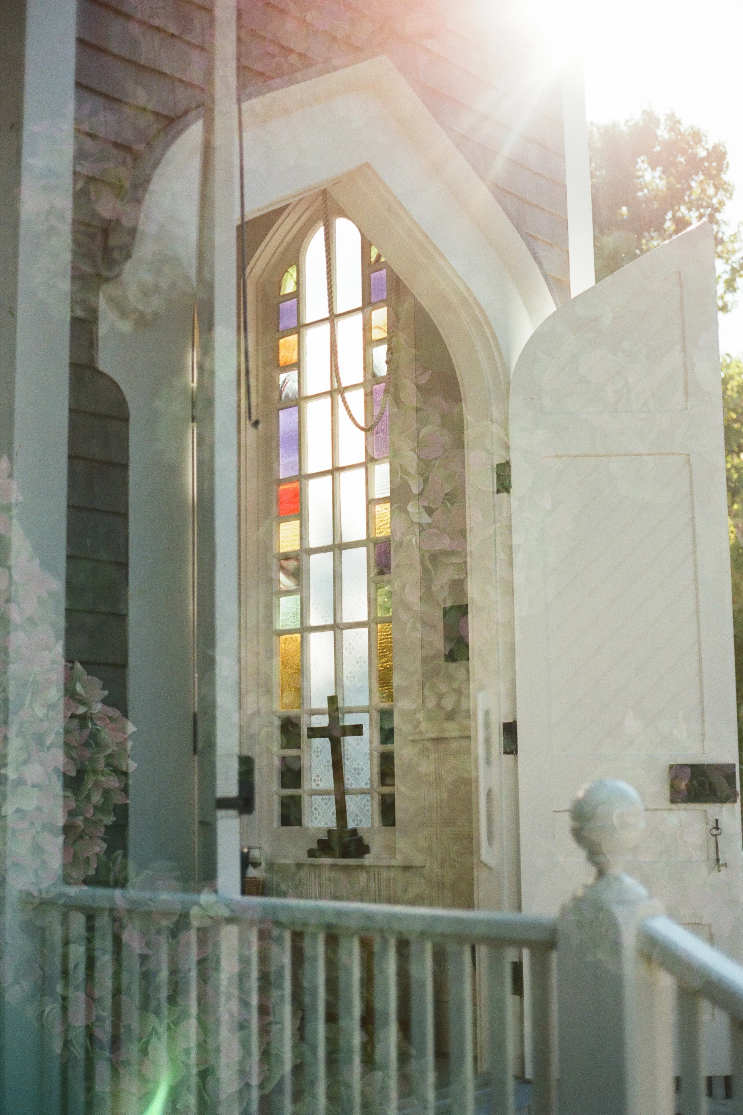 Sunlight streaming through a stained glass window in a church, with an open door and a wooden cross on the window ledge.