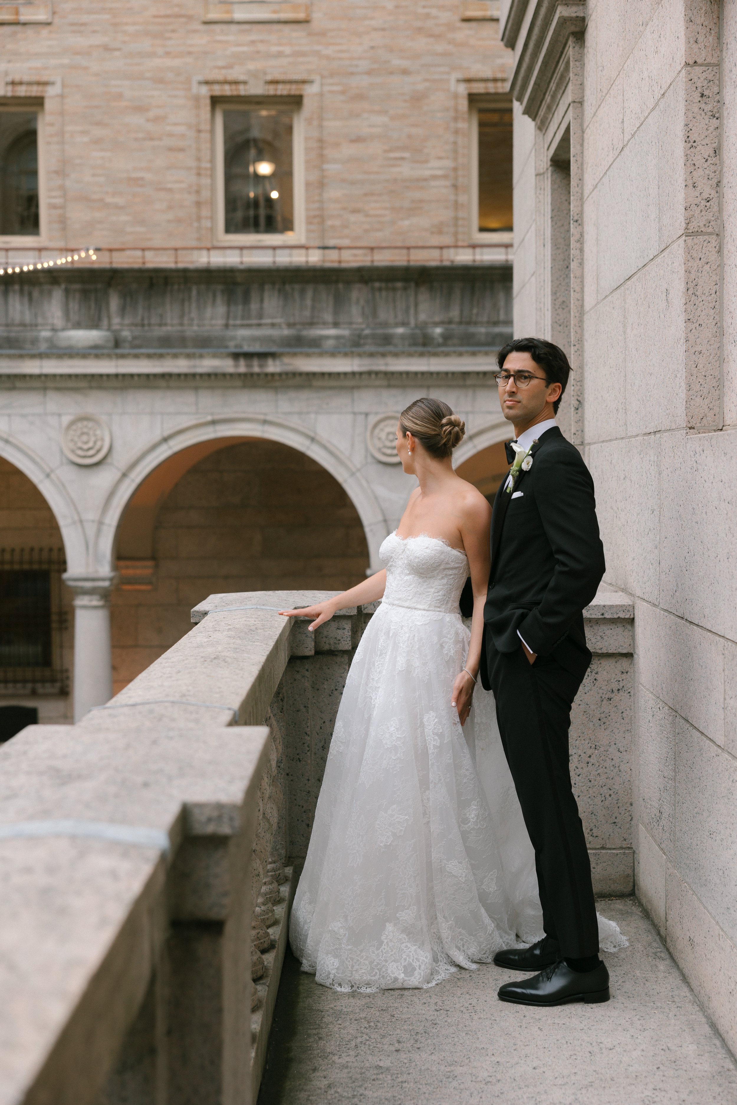 A bride in a white lace wedding dress and a groom in a black tuxedo pose outdoors on a stone balcony with arched architecture behind them.