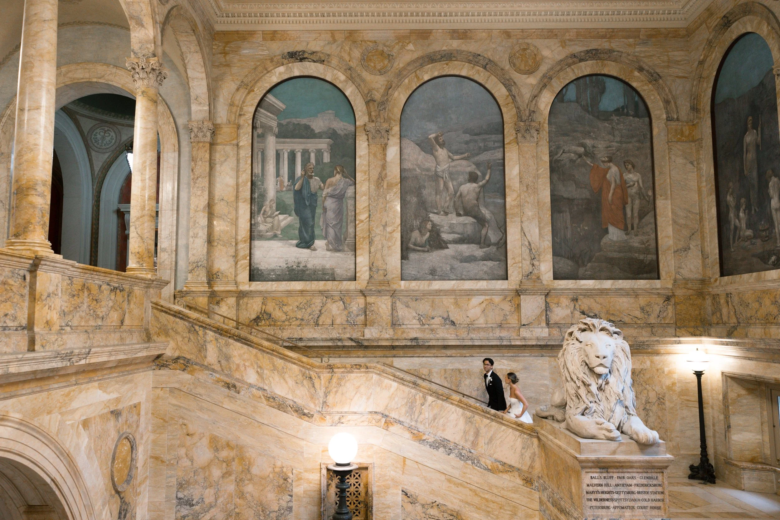 A bride and groom in wedding attire walking up a grand marble staircase inside a building with classical paintings and sculptures, including a large lion statue at the bottom of the stairs.