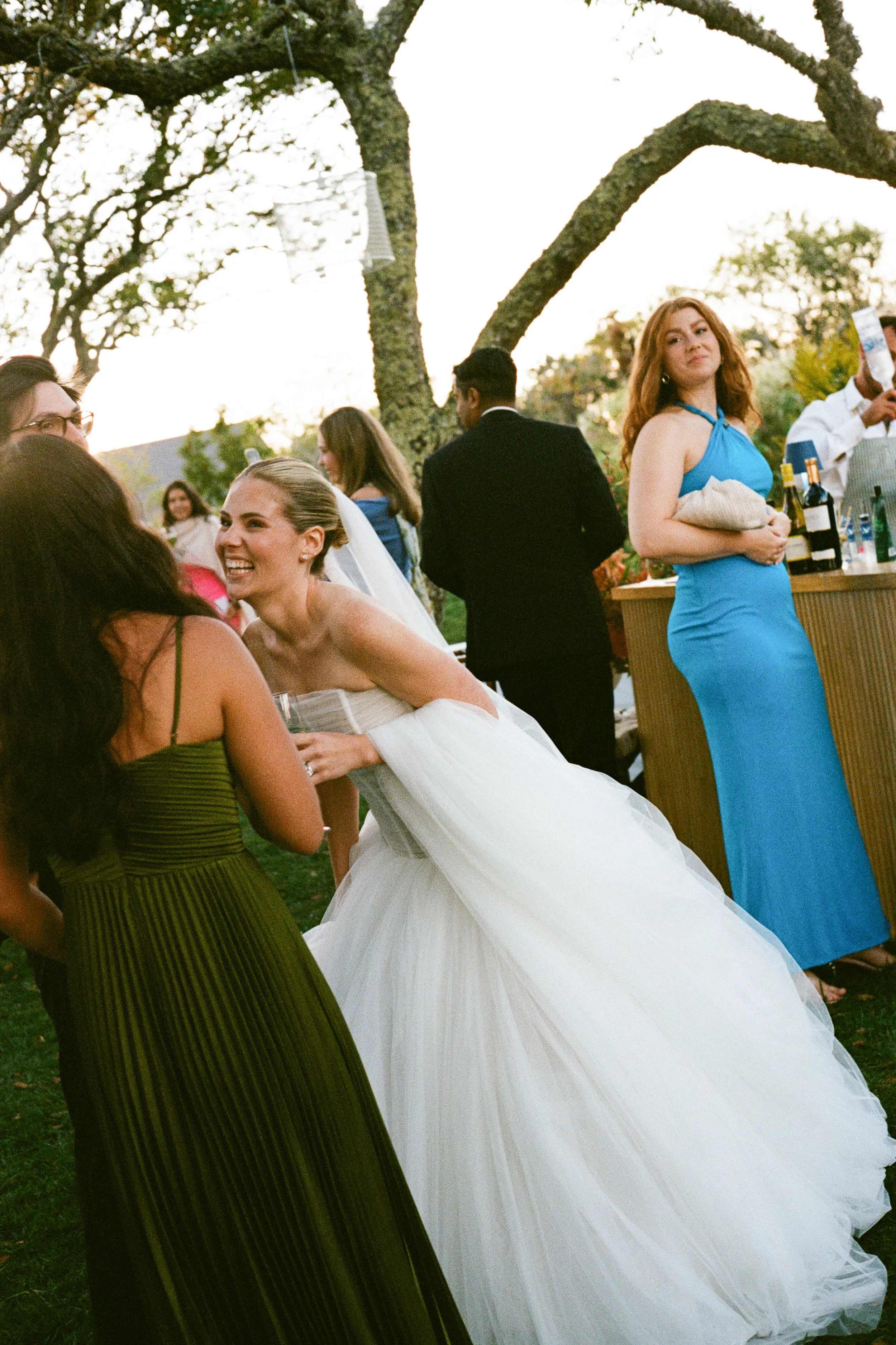 Bride laughing and talking with guests at an outdoor wedding reception under trees, with a bar area in the background.
