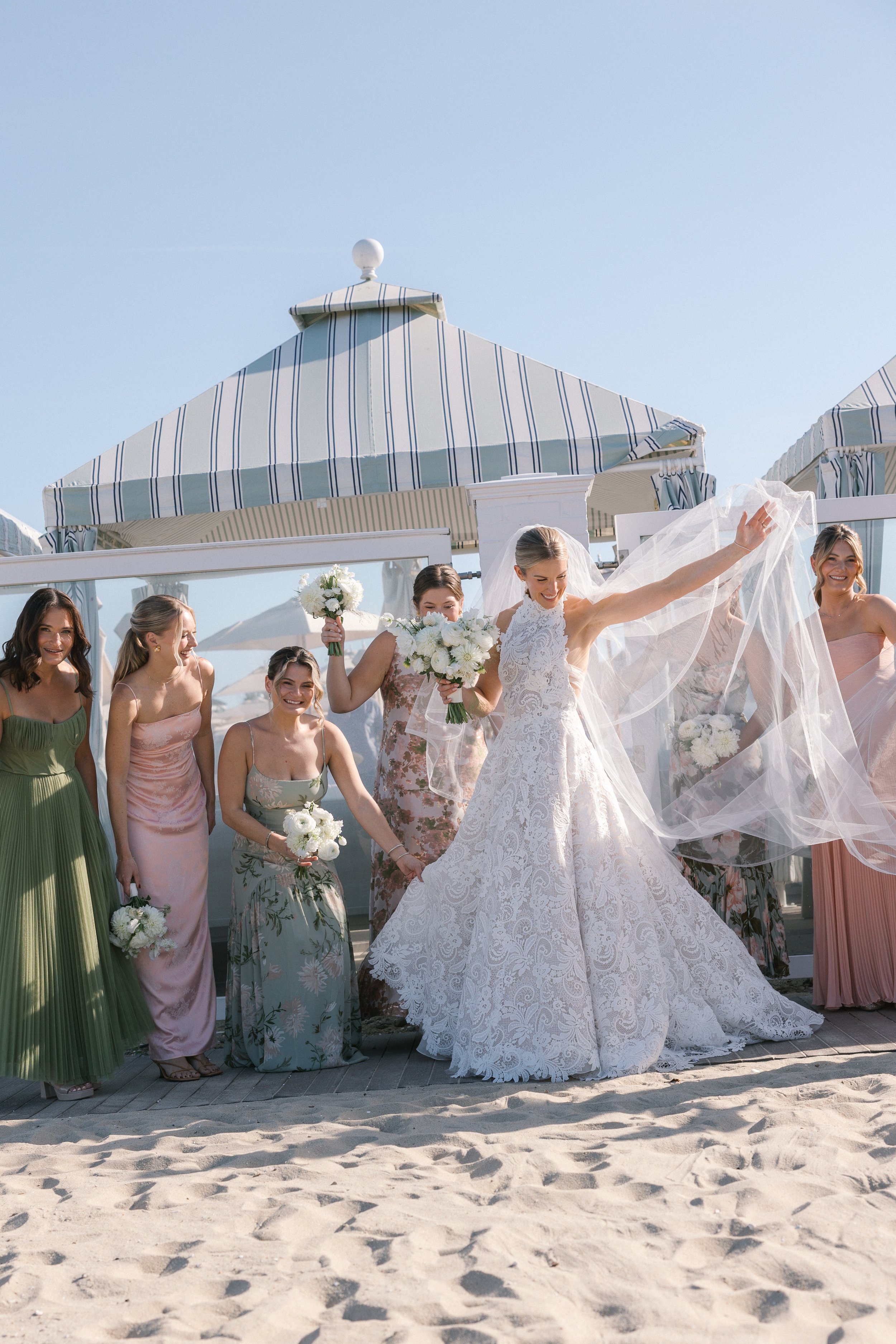 A bride and her bridesmaids celebrating on the beach, with the bride in a white lace wedding dress and the bridesmaids in pastel-colored dresses, holding bouquets, under a large beach umbrella.