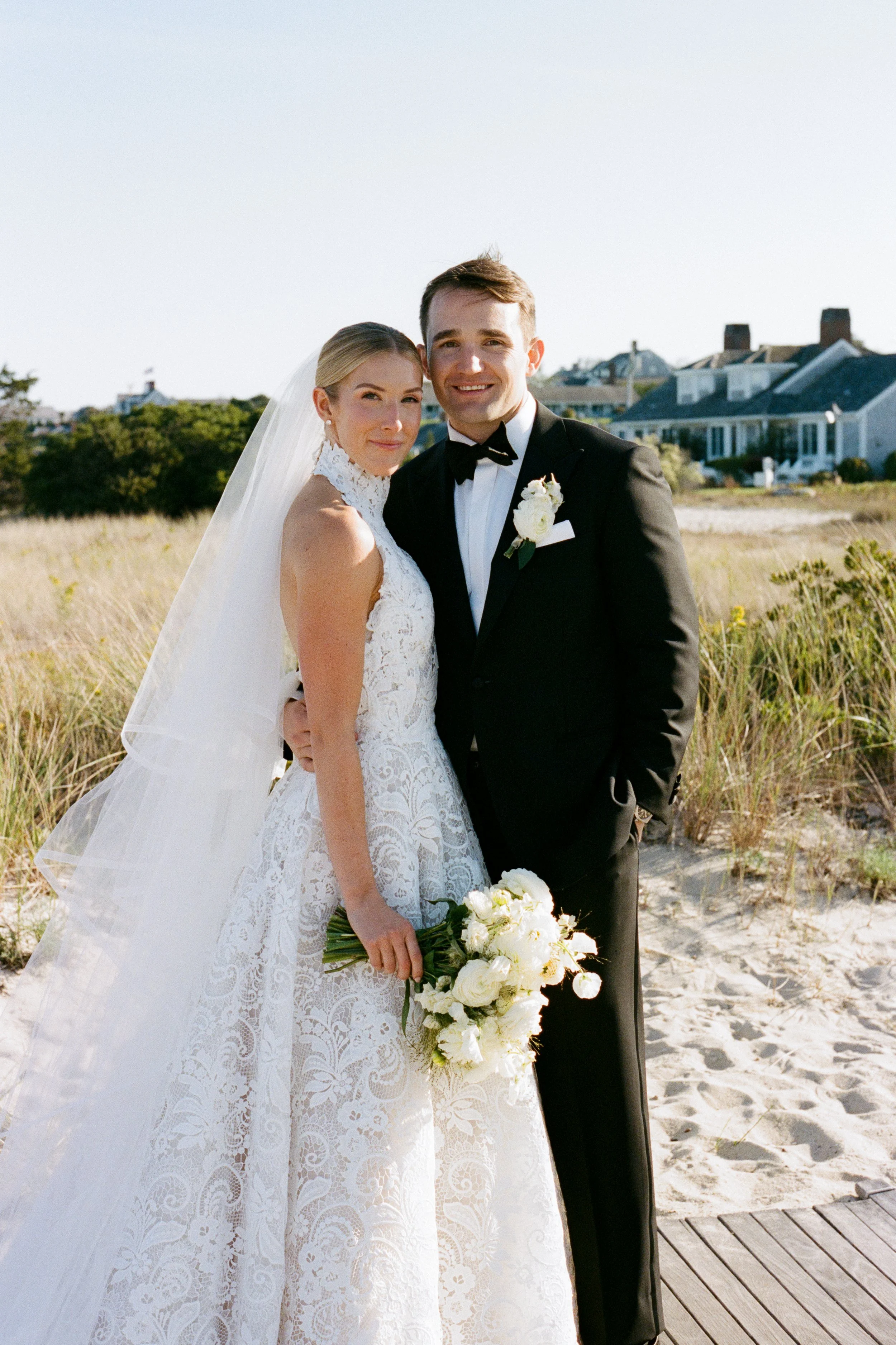 A bride and groom standing together outdoors on a sunny day, with bridal attire, a bouquet of white flowers, and beach houses in the background.
