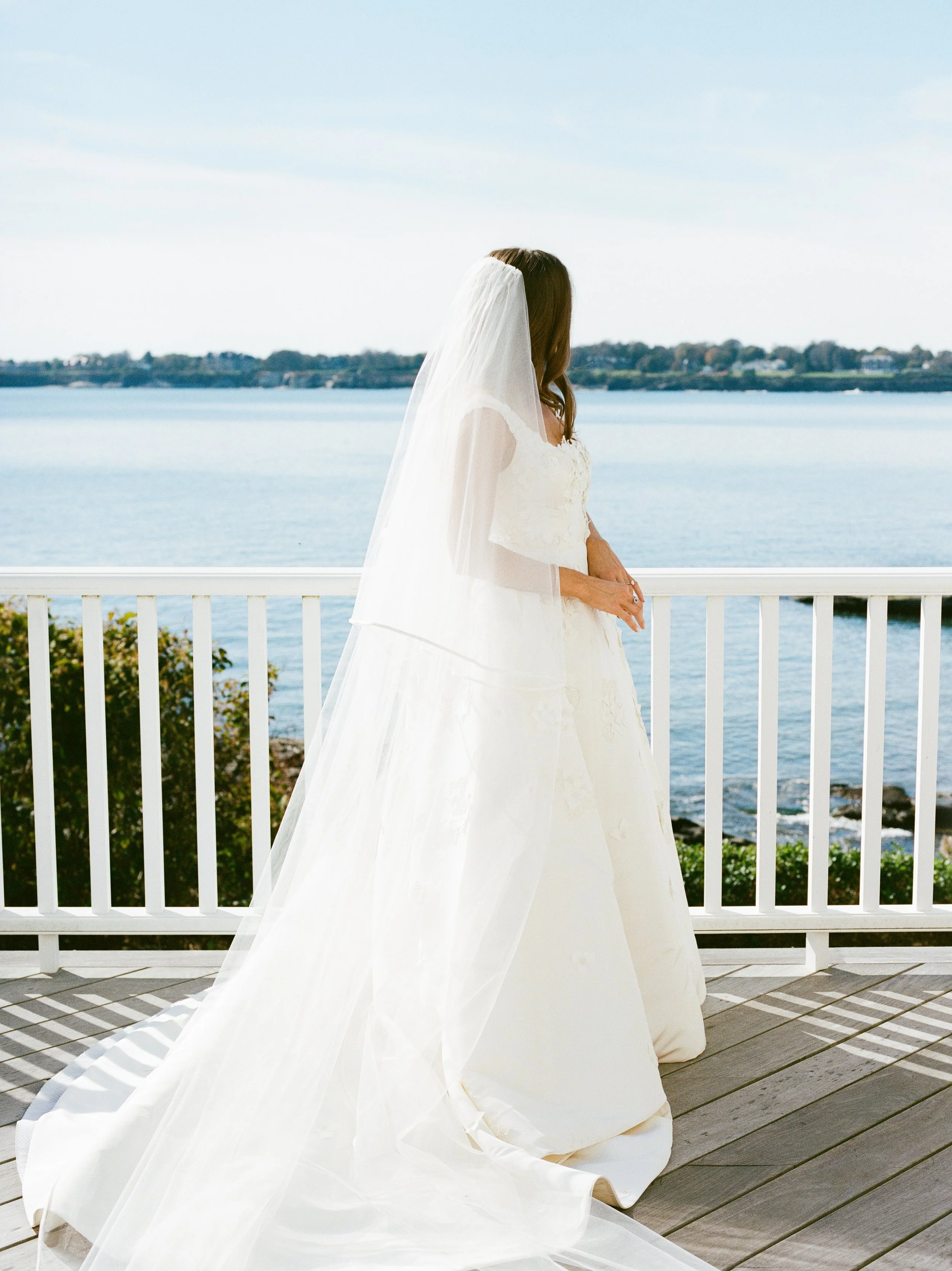 Bride in a white wedding dress and veil standing on a balcony overlooking a lake or ocean, with trees and land in the distance.