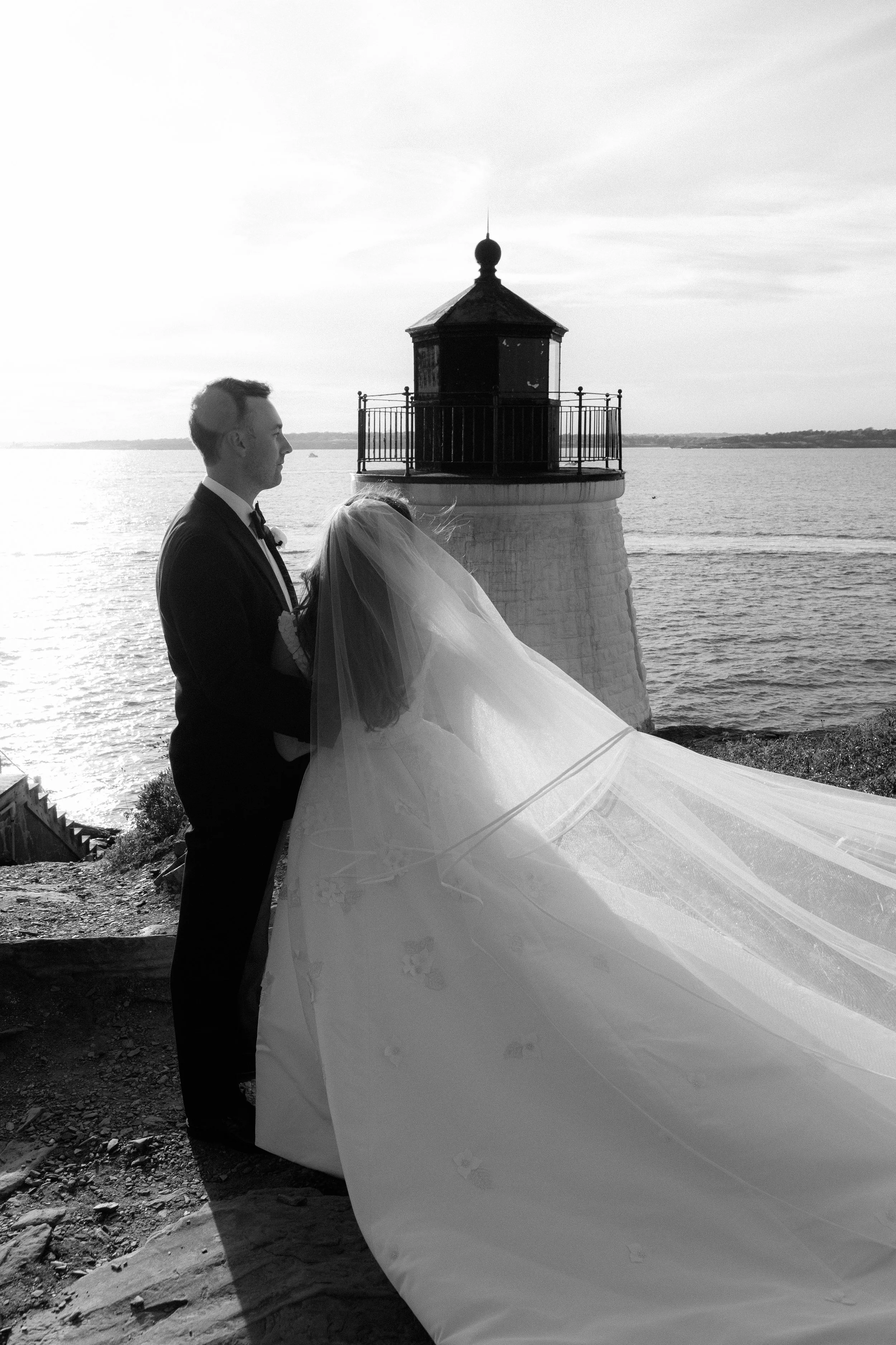 A black and white photo of a bride and groom standing near a lighthouse by the water. The groom is in a tuxedo, holding the bride, who is in a wedding dress with a long veil.