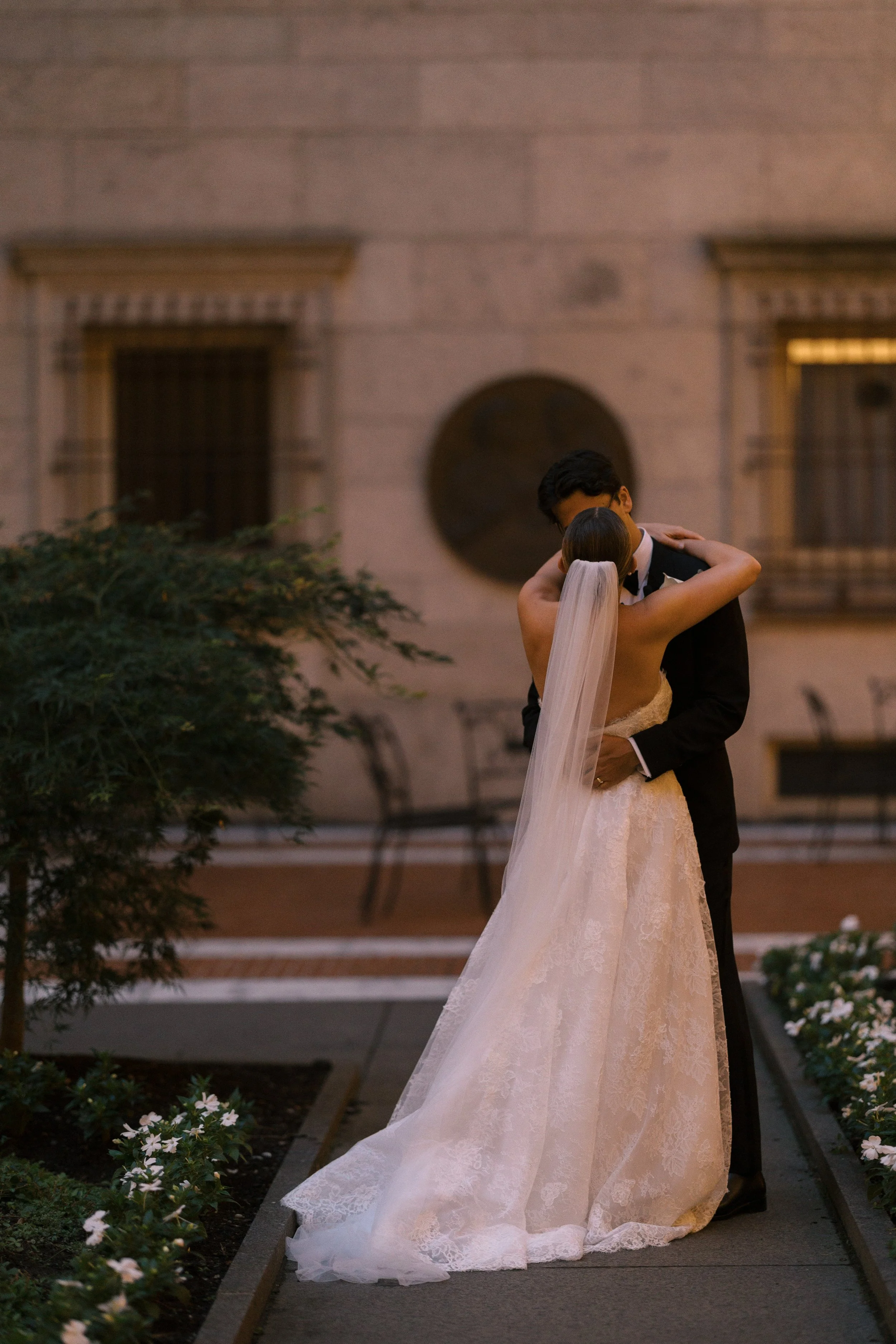 Bride and groom embracing outdoors at sunset, with the bride in a lace wedding gown and veil, and the groom in a suit, against a historic building background.