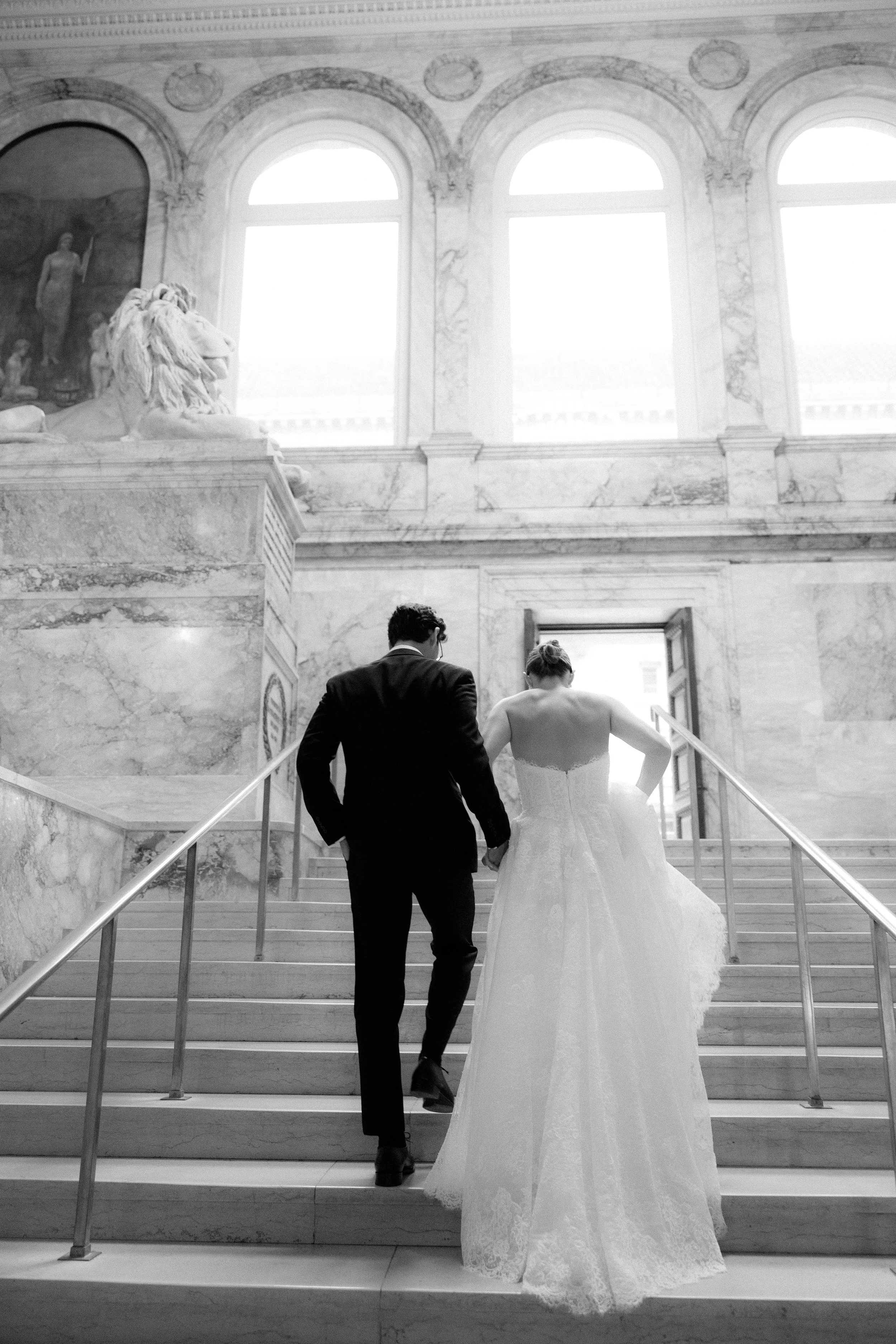A bride and groom ascending a marble staircase inside a grand, historic building with large arched windows and classical sculptures, including a lion statue.