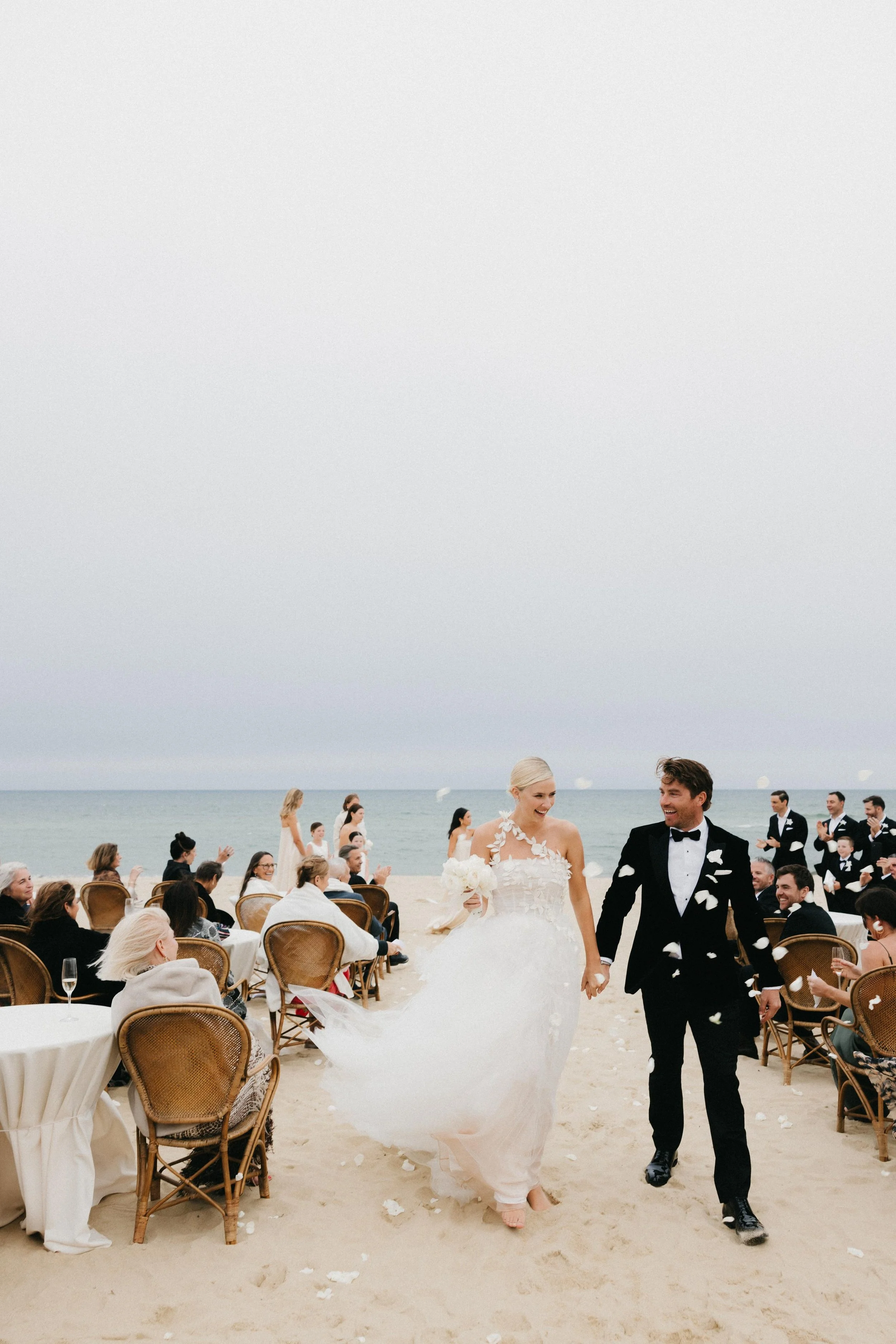 A bride and groom walking hand in hand on a beach during their wedding ceremony, surrounded by seated guests and bridesmaids, with the ocean and cloudy sky in the background.