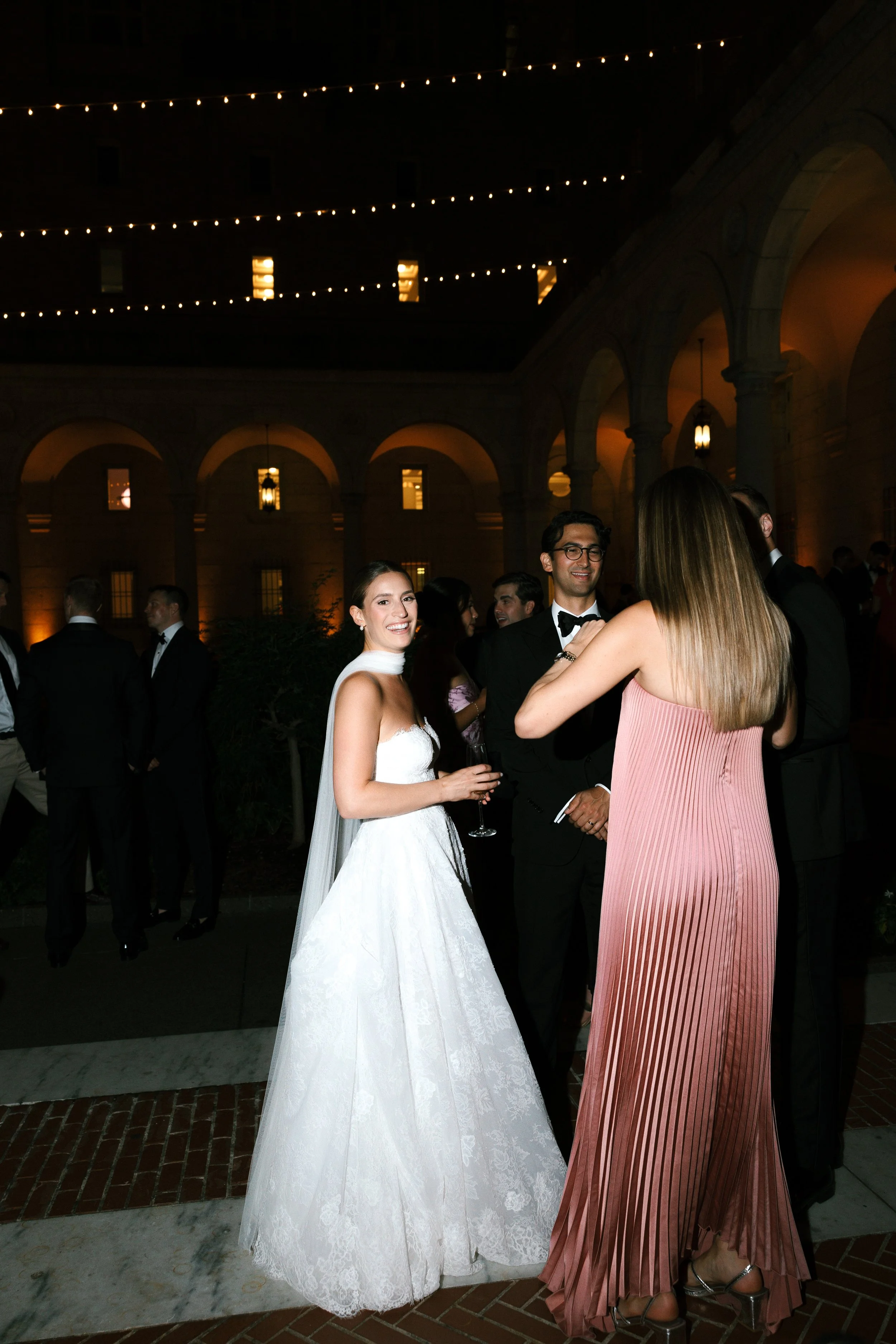 A group of people attending a formal event or wedding reception outdoors at night, with string lights and arched architectural features in the background. In the foreground, a woman in a white wedding dress is smiling, holding a glass, while talking 