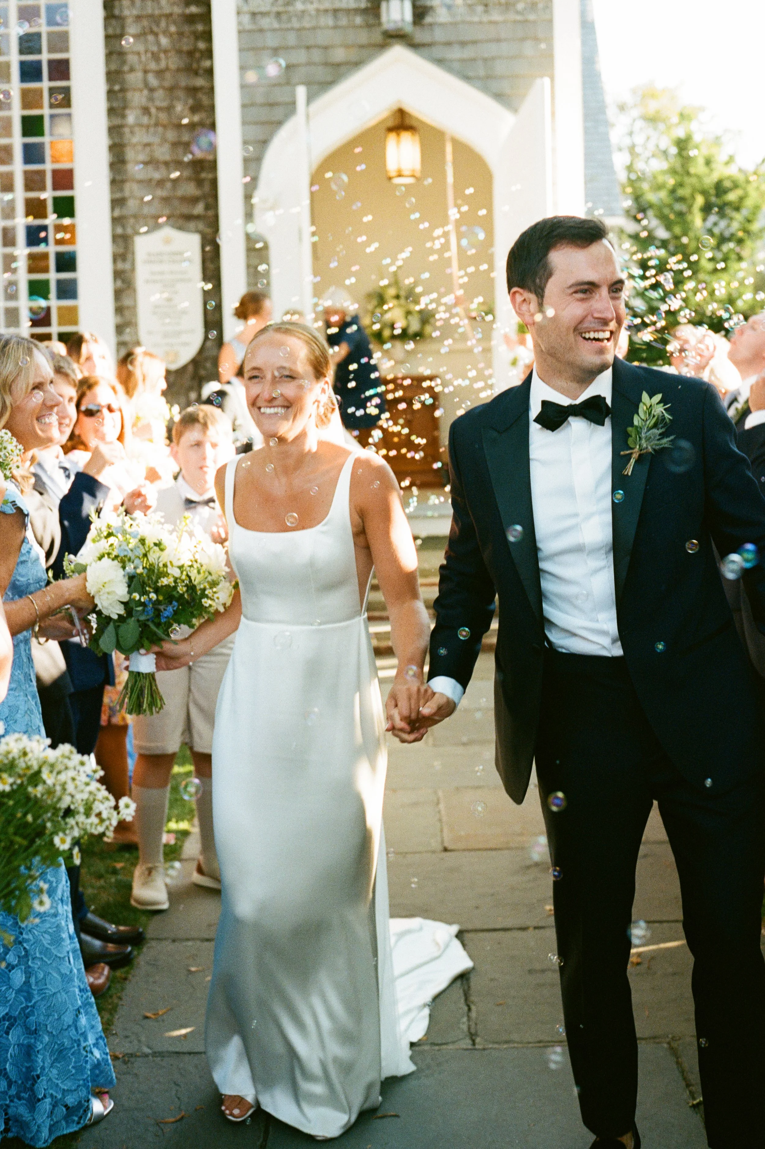 Bride and groom holding hands, walking out of a church celebrating their wedding with guests throwing bubbles.
