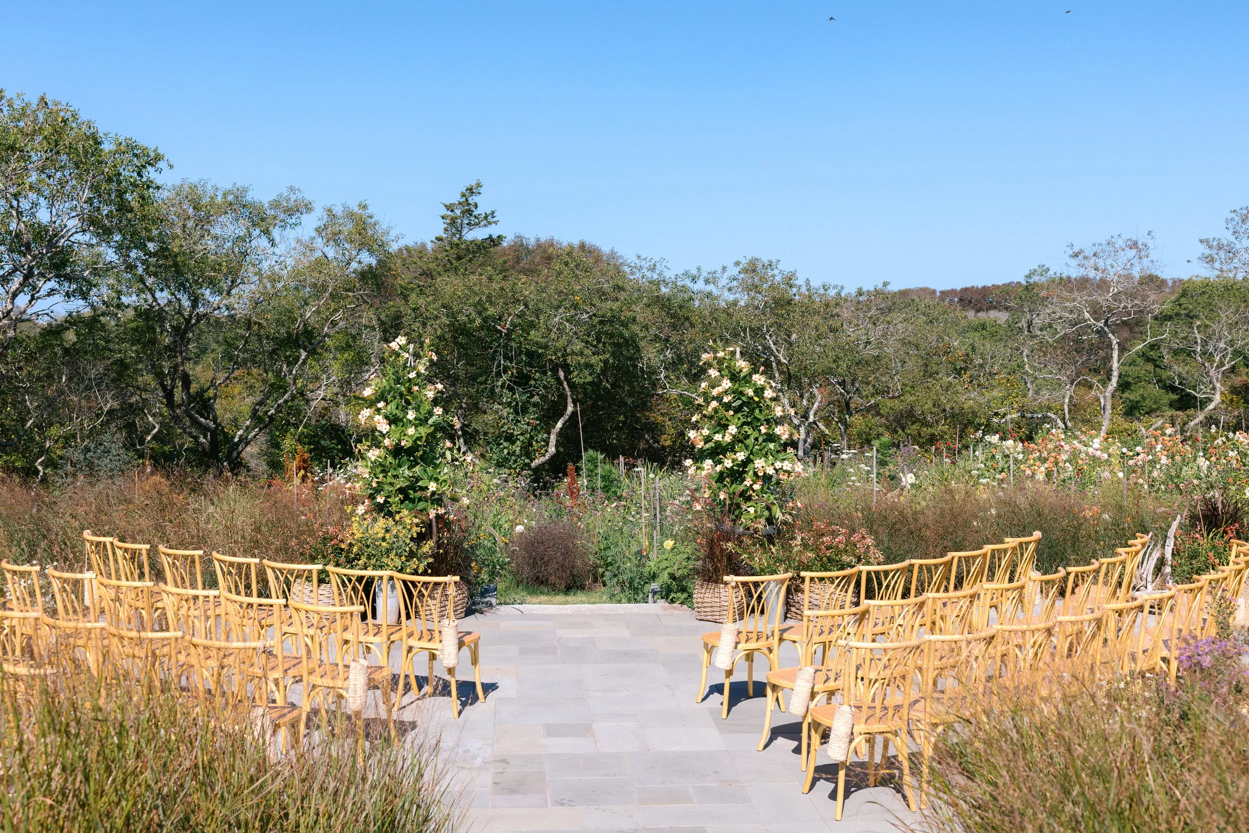 Outdoor wedding or event ceremony setup with wooden chairs arranged in rows facing an altar or focal point, surrounded by lush greenery and flowering plants, under a clear blue sky.