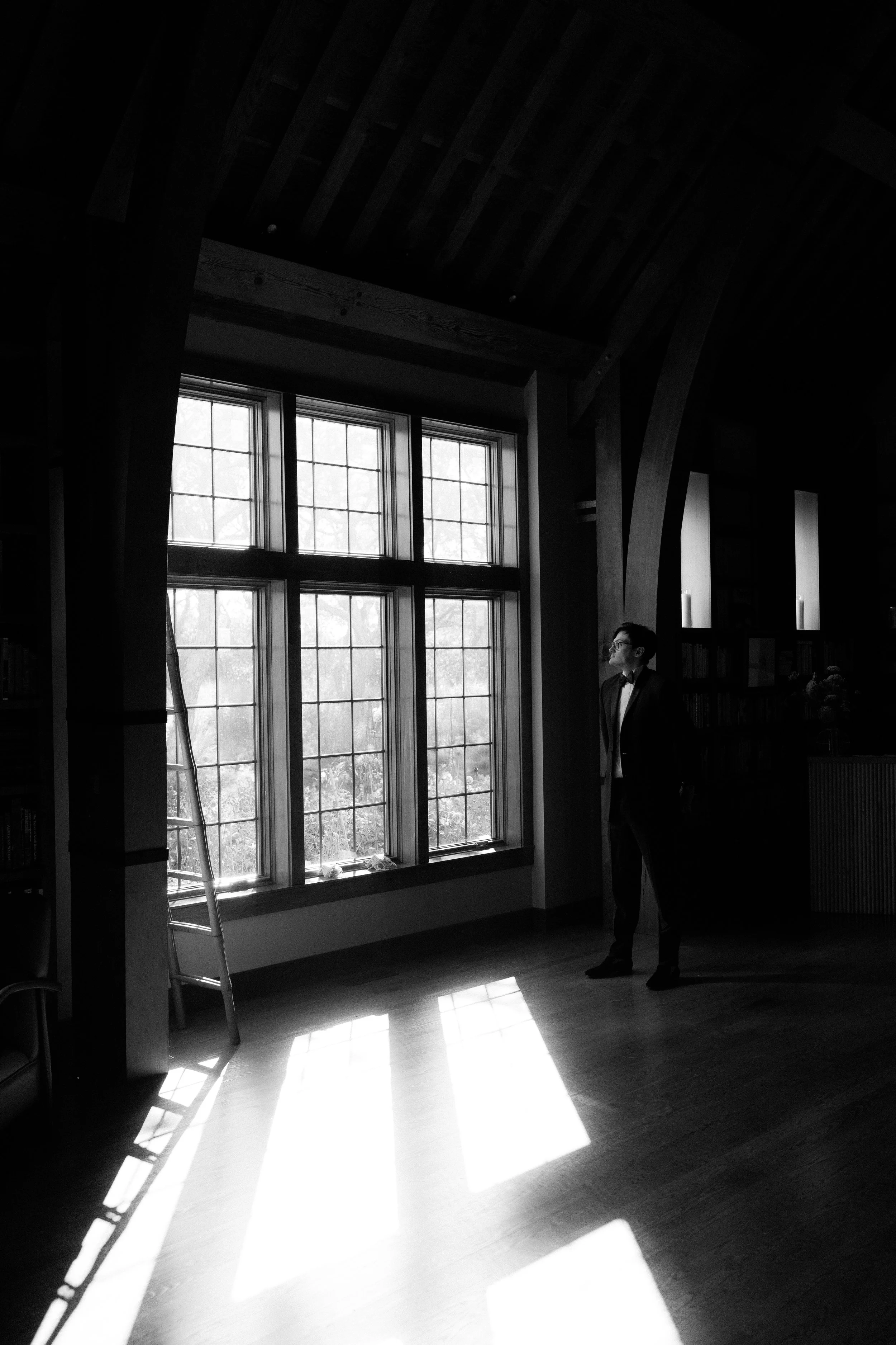 A man dressed in a tuxedo standing in a dimly lit room, looking out the large window with sunlight casting rectangular light patterns on the wooden floor.