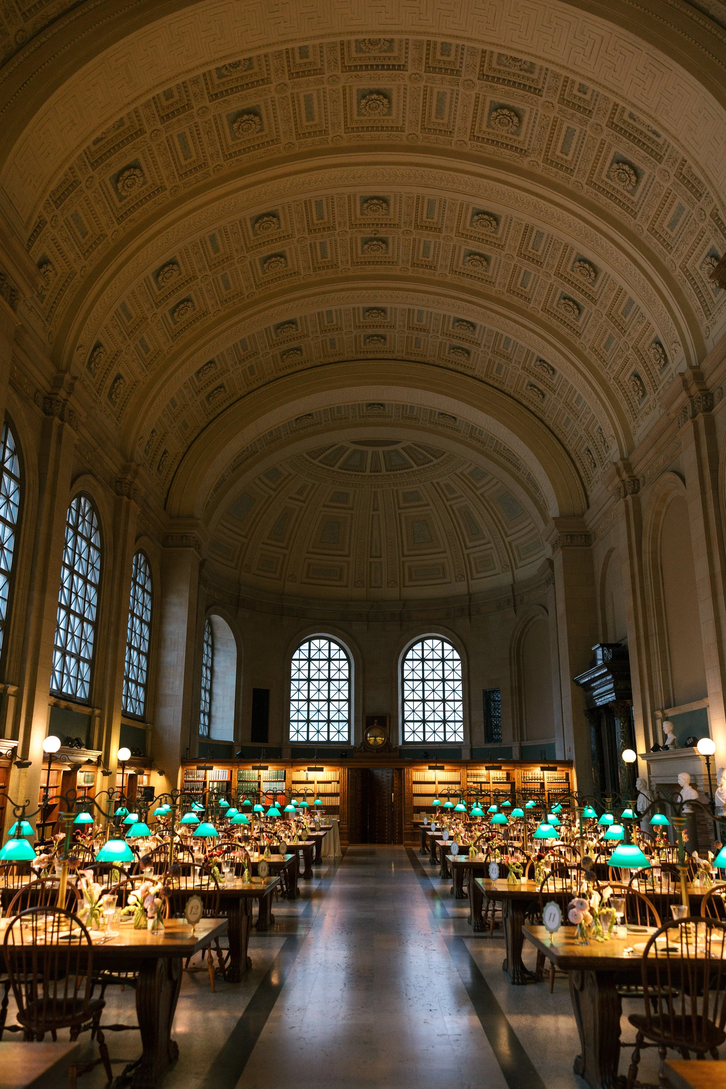 Elegant grand hall with high, decorated ceiling, large arched windows, and a long central aisle. Rows of tables with green-shaded lamps, floral centerpieces, bookshelves on the back wall, and classical bust sculptures on the right side.