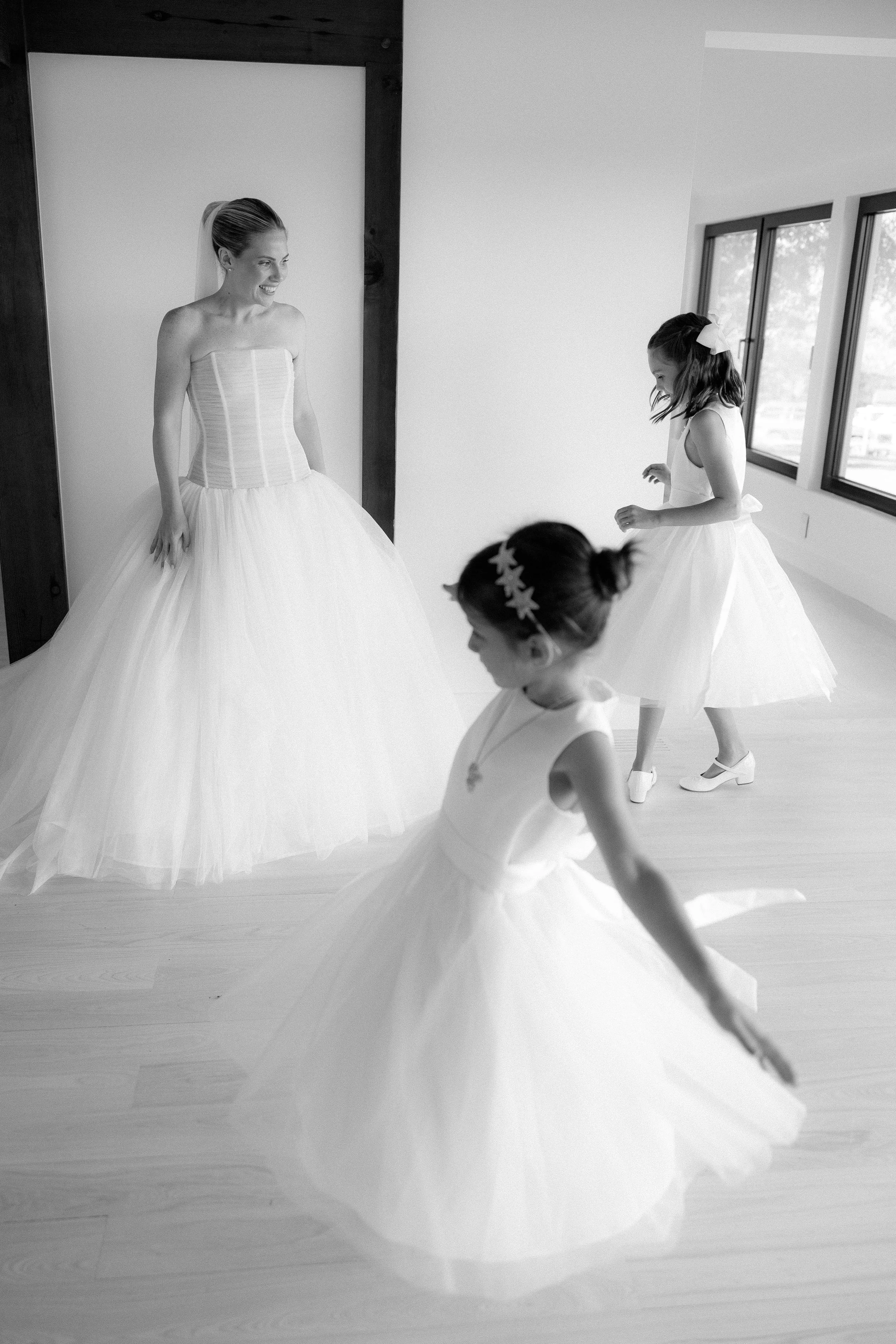 A bride and two young girls in white dresses, possibly flower girls, dancing and smiling during a wedding preparation in a room with large windows.