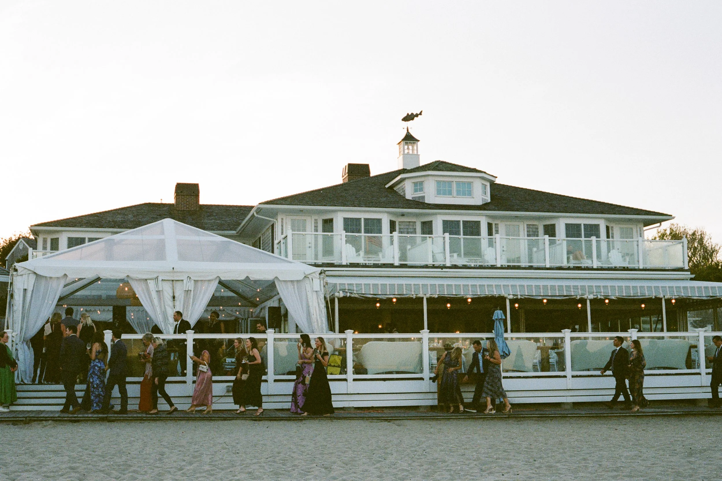 Guests gathering outside a white, two-story house with a deck, used as a venue for an event, on a sandy beach during sunset.