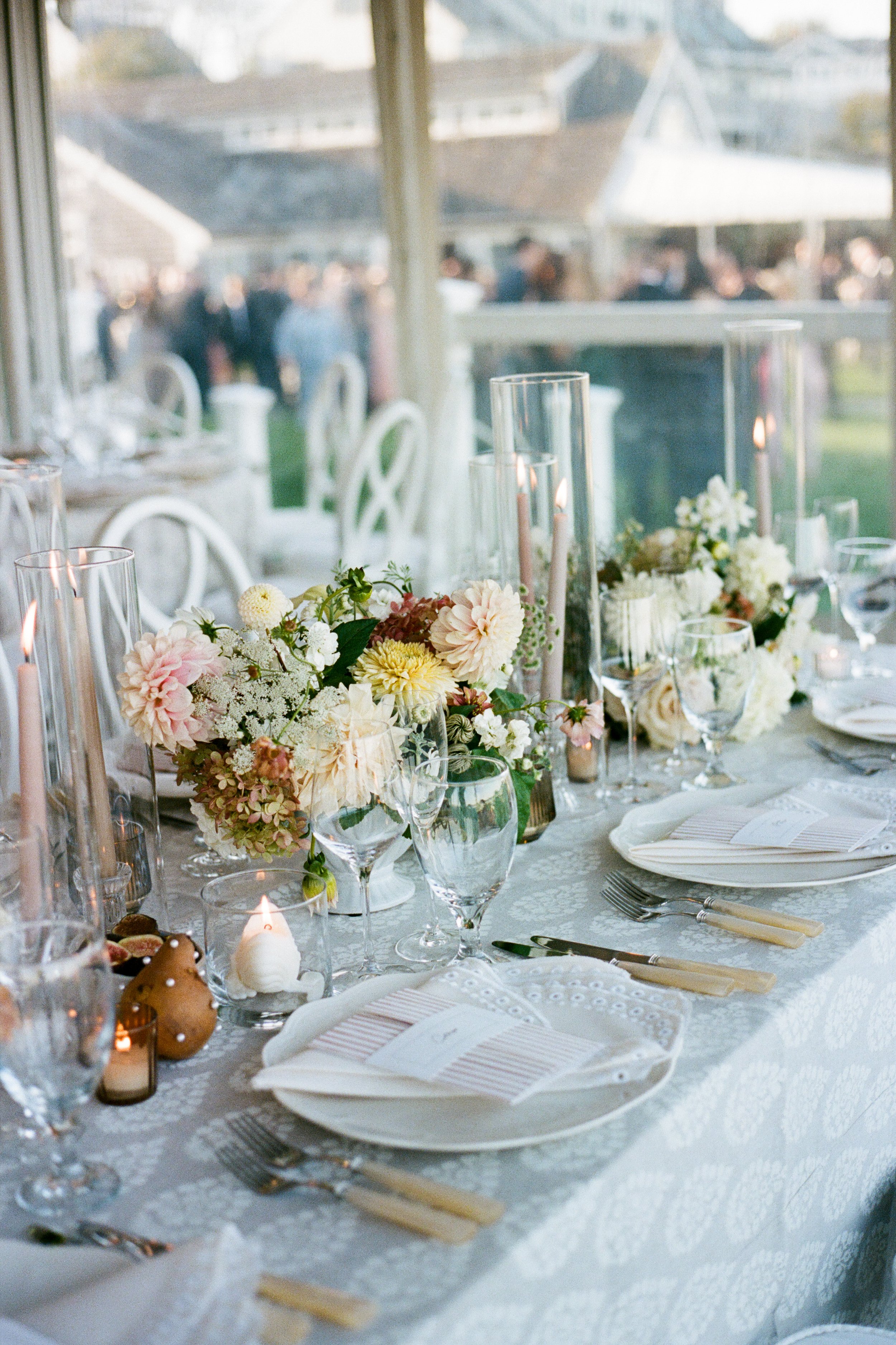 Elegant wedding or event table setting with floral centerpieces, tall glass candle holders, plates, cutlery, and glasses, with a window view of a gathering outside.