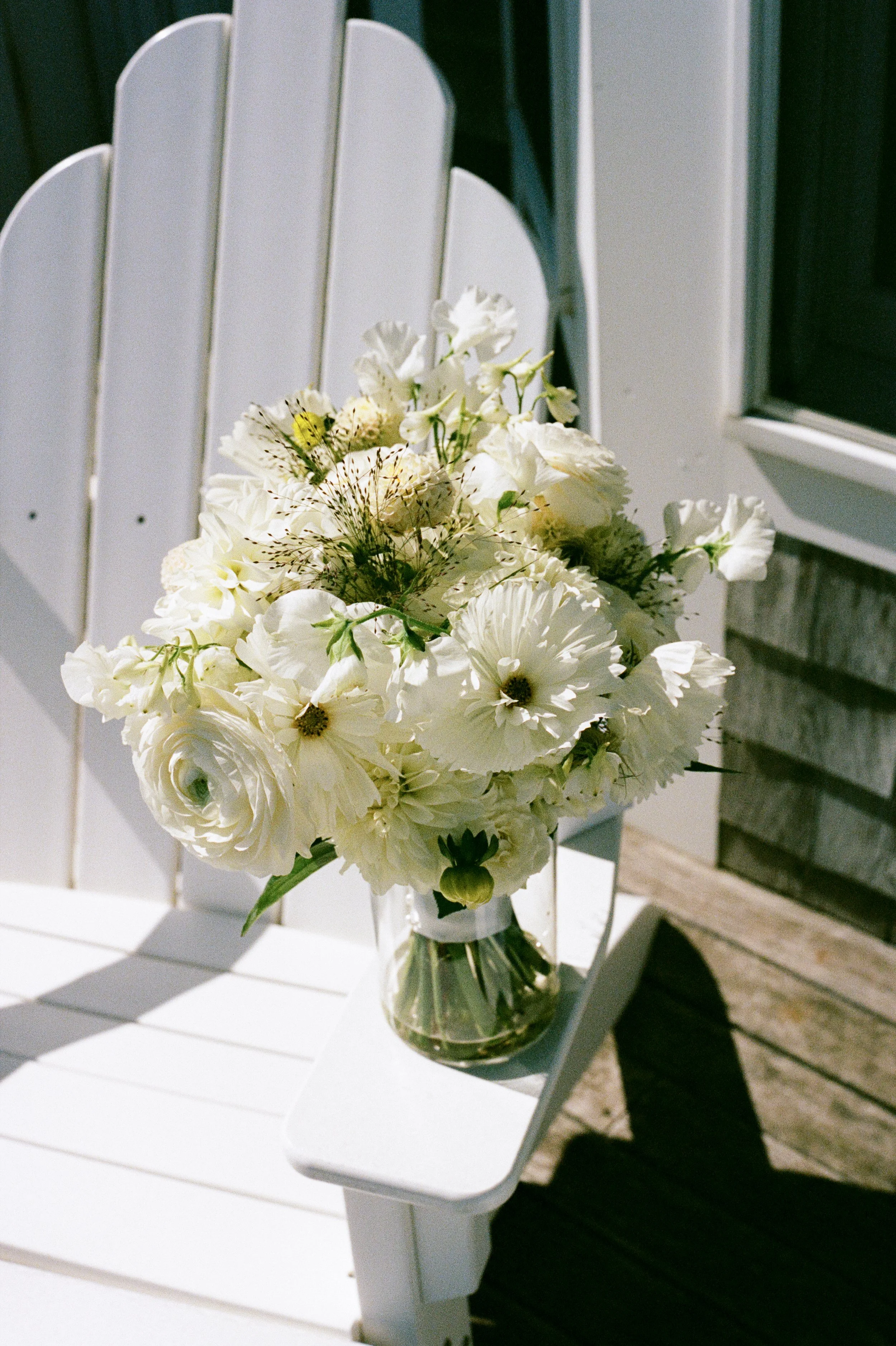 A glass vase filled with white flowers, placed on a white chair outdoors near a building.