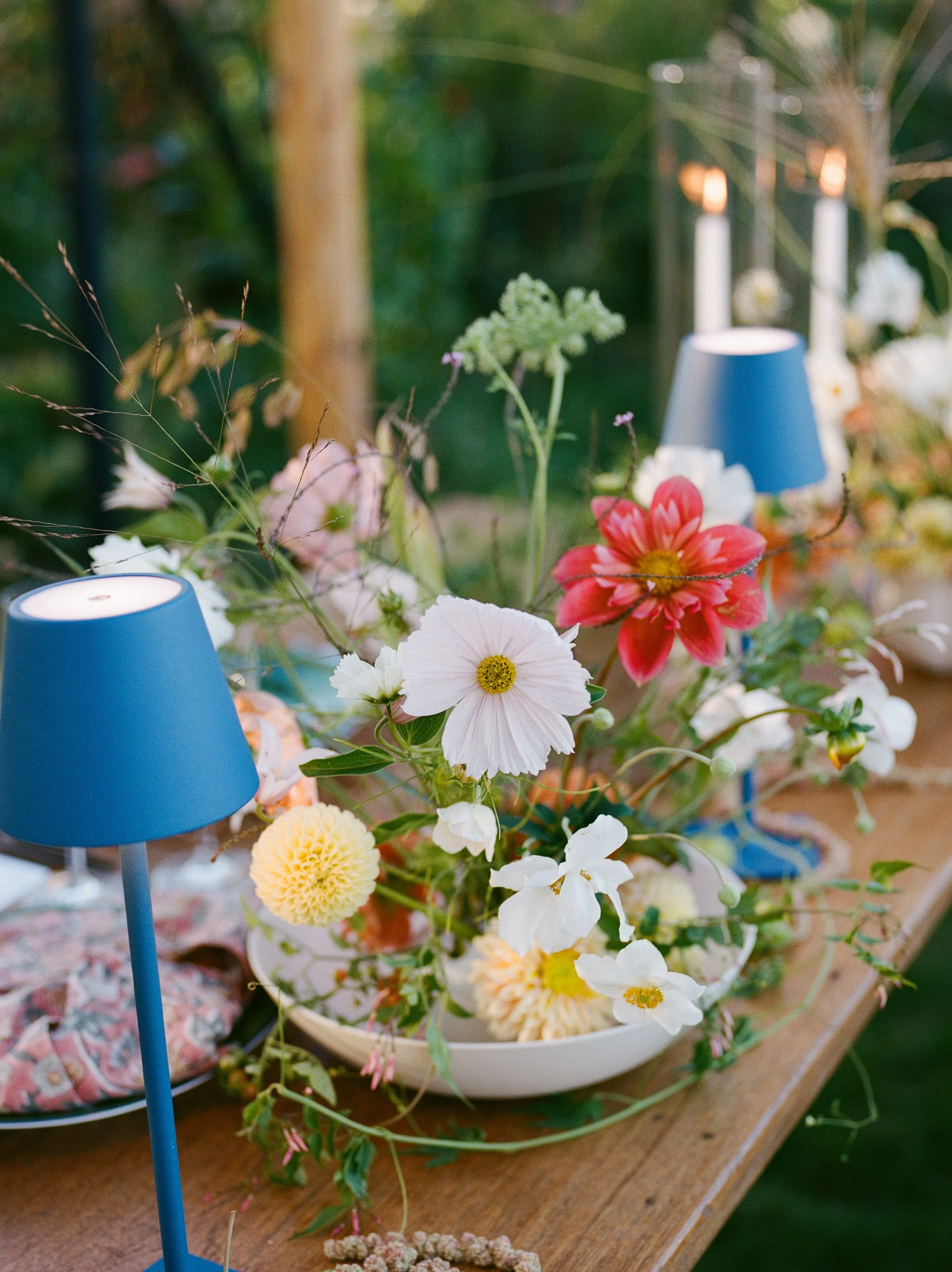 A wooden table decorated with a floral arrangement featuring various colorful flowers, two blue candle holders, one in focus, and a patterned cloth. The background is blurred with greenery.