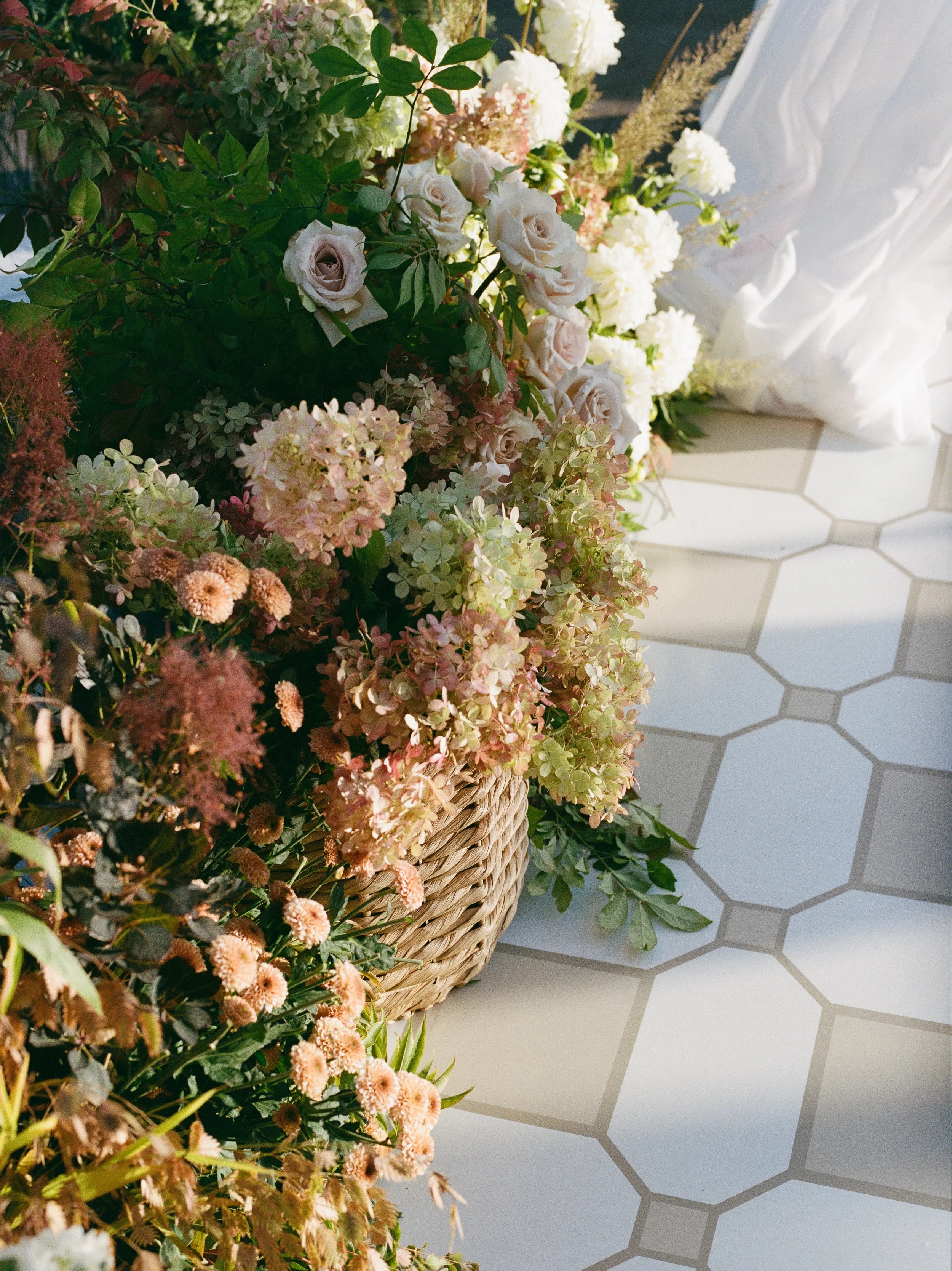A woven basket filled with various light-colored flowers, including roses and hydrangeas, placed on a white tiled surface with sunlight casting shadows.
