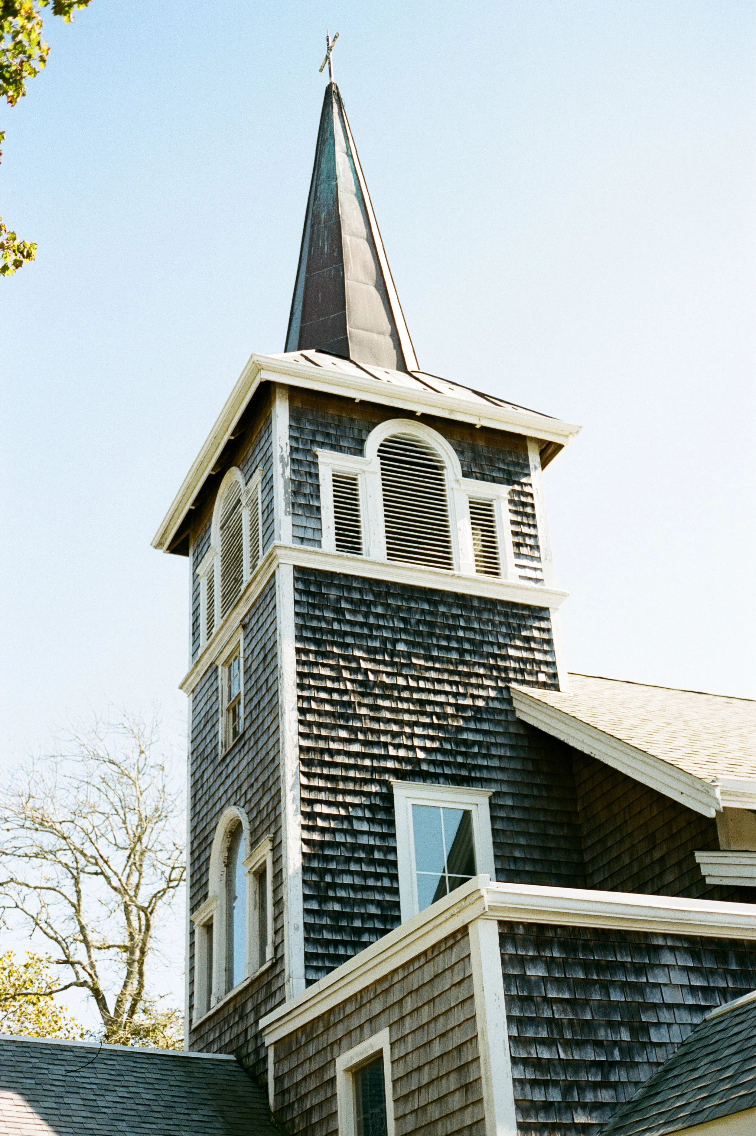 A close-up of a church steeple with a cross on top, above a building with weathered black wooden shingles and white window frames, under a clear sky.