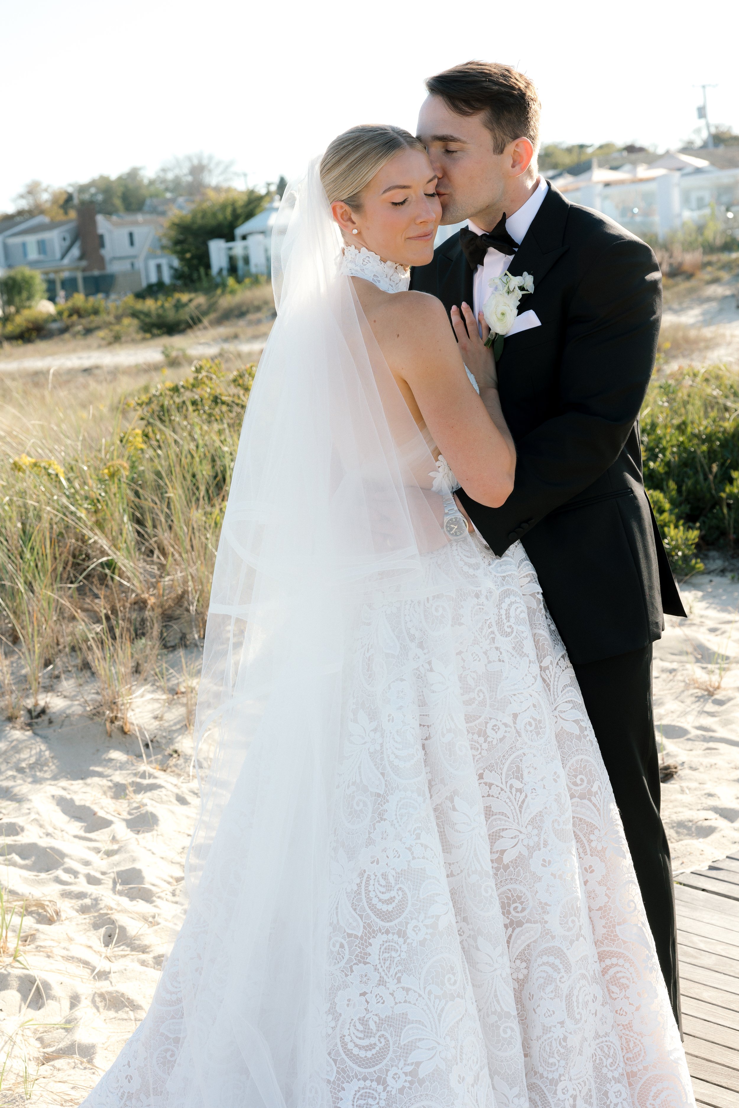 A bride and groom in wedding attire embrace on a sandy beach with houses and vegetation in the background.