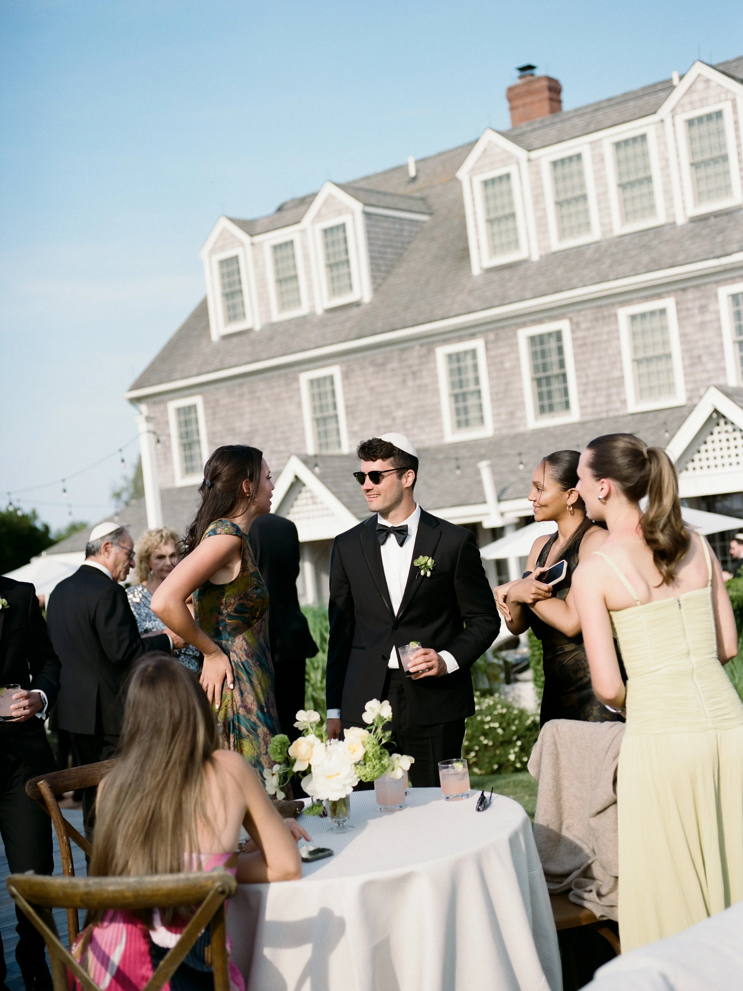 People dressed in formal attire socializing outdoors at a gathering, with a large house in the background.