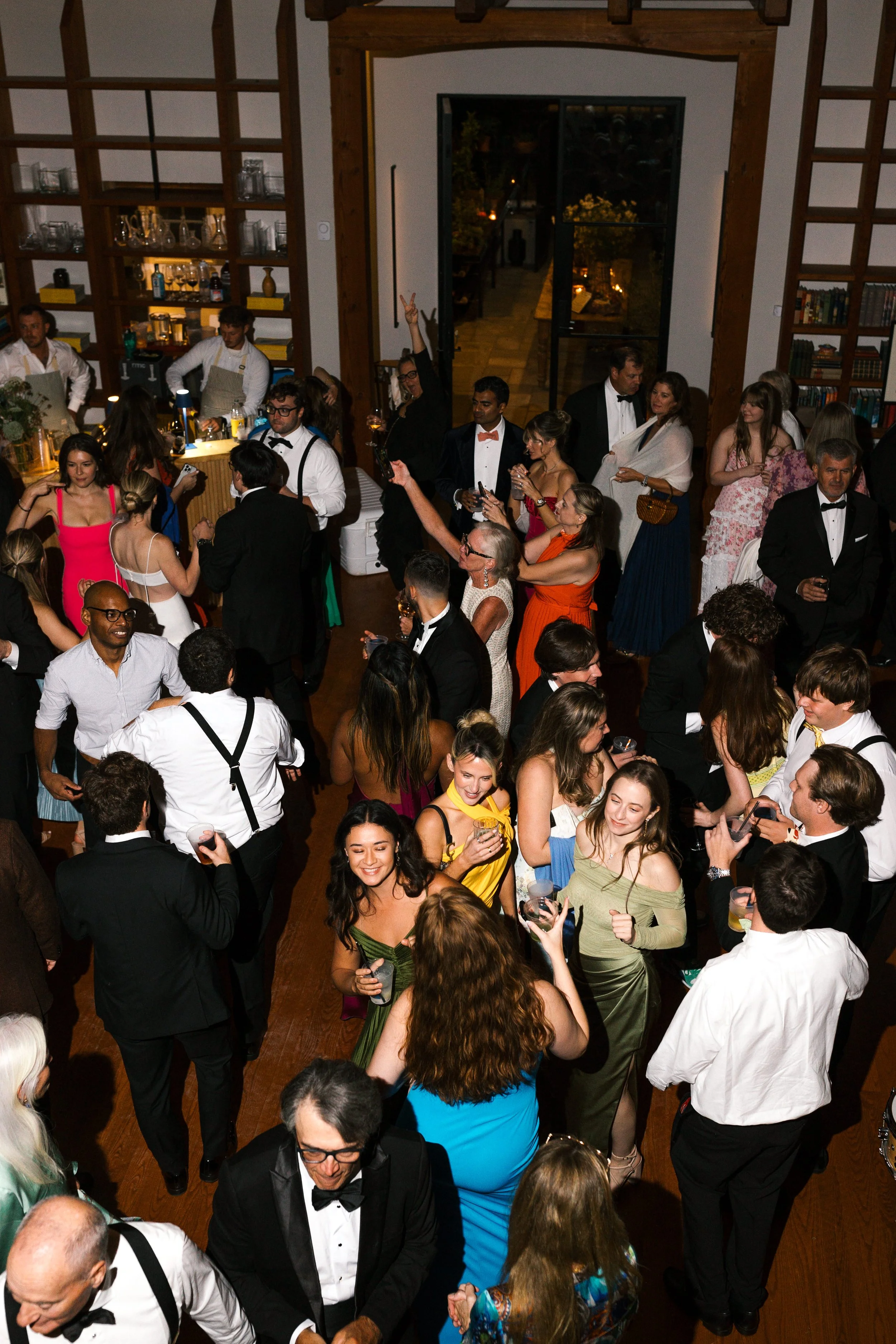 People dancing and socializing at a party in a large, well-lit room with wooden flooring and bookshelves, some holding drinks, dressed in formal attire.