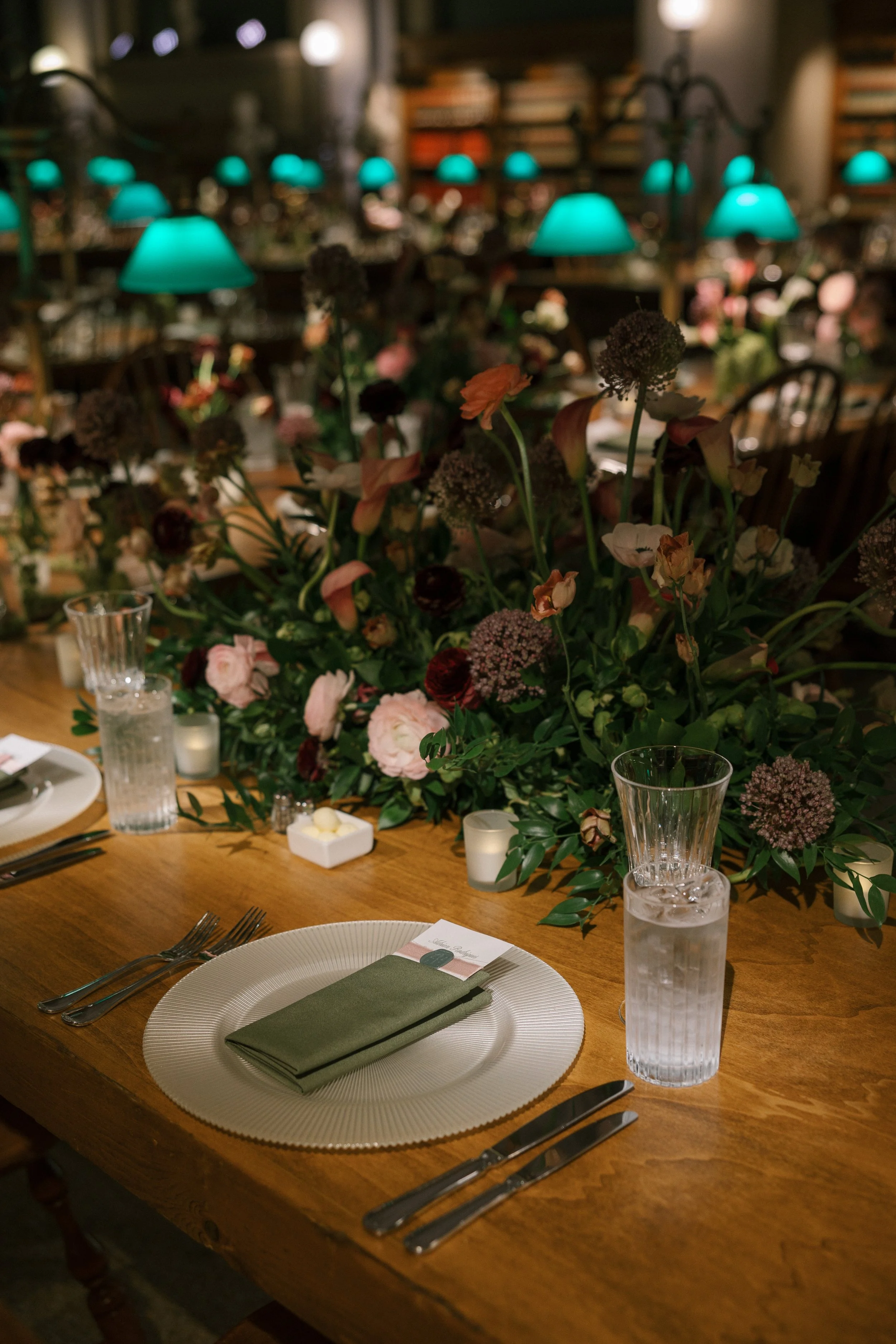 A formal dining table decorated with a large floral centerpiece, candles, glasses of water, and place settings with a white plate, green napkin, and silverware.