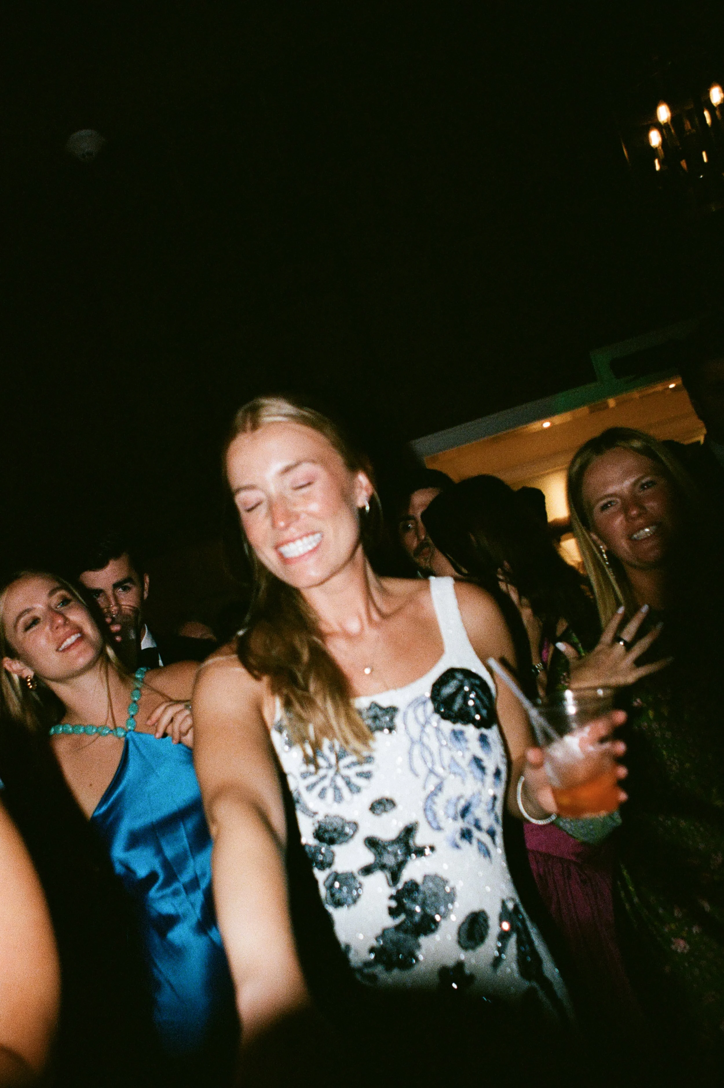 A group of people dancing at a party. A woman in the foreground is smiling with eyes closed, holding a drink, wearing a white dress with black floral patterns. Others around her are also enjoying the event.