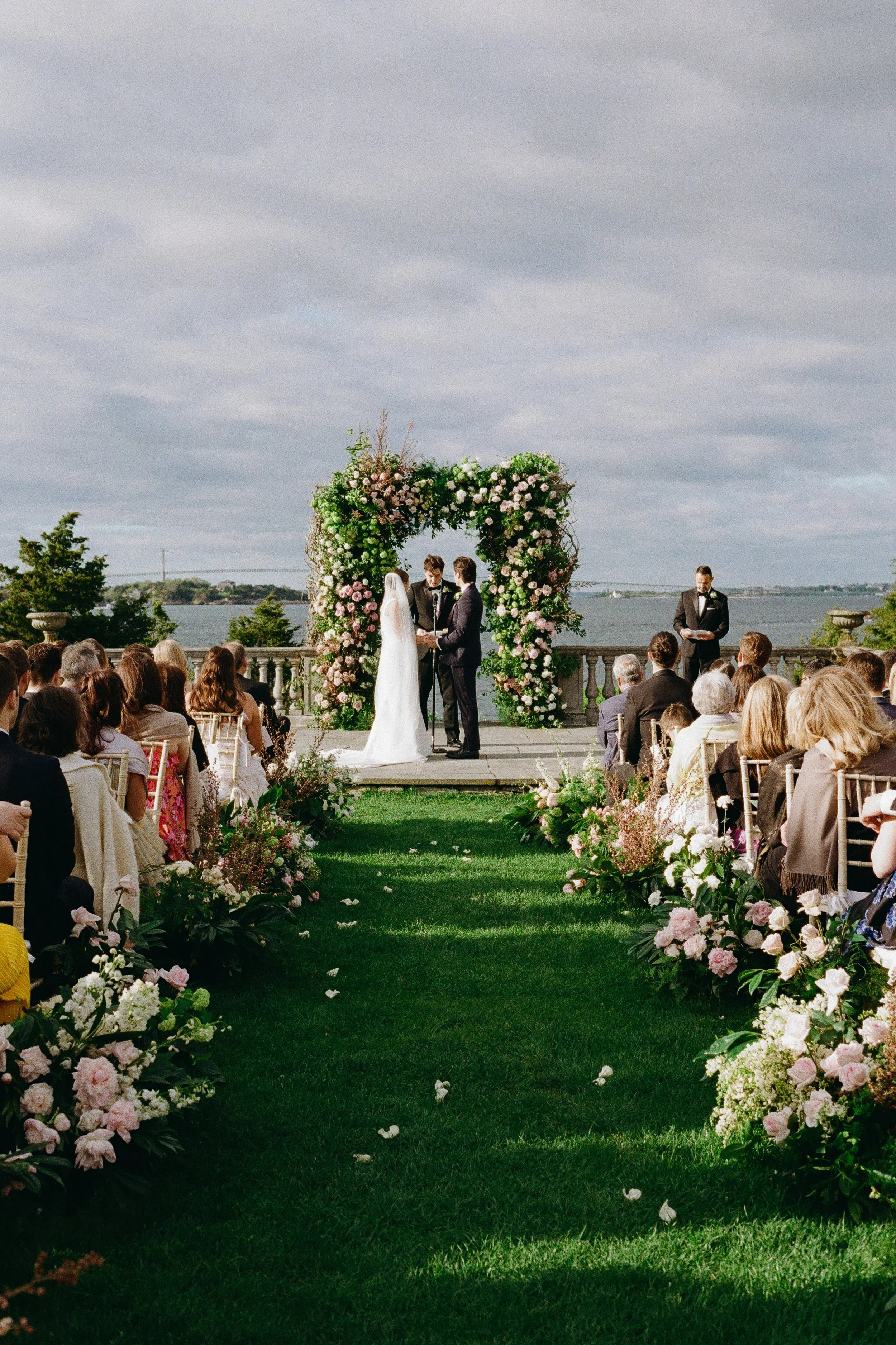A wedding ceremony outdoors with a couple exchanging vows under a floral arch by a body of water, with guests seated on either side of a green grass aisle.