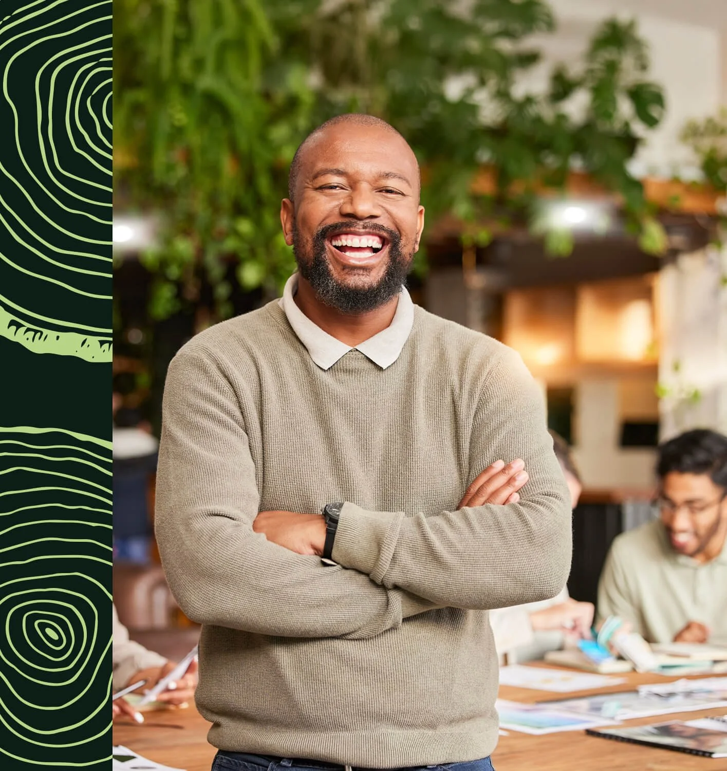 Homme souriant avec les bras croisés dans un espace intérieur paisible et verdoyant, avec d'autres personnes en arrière-plan.