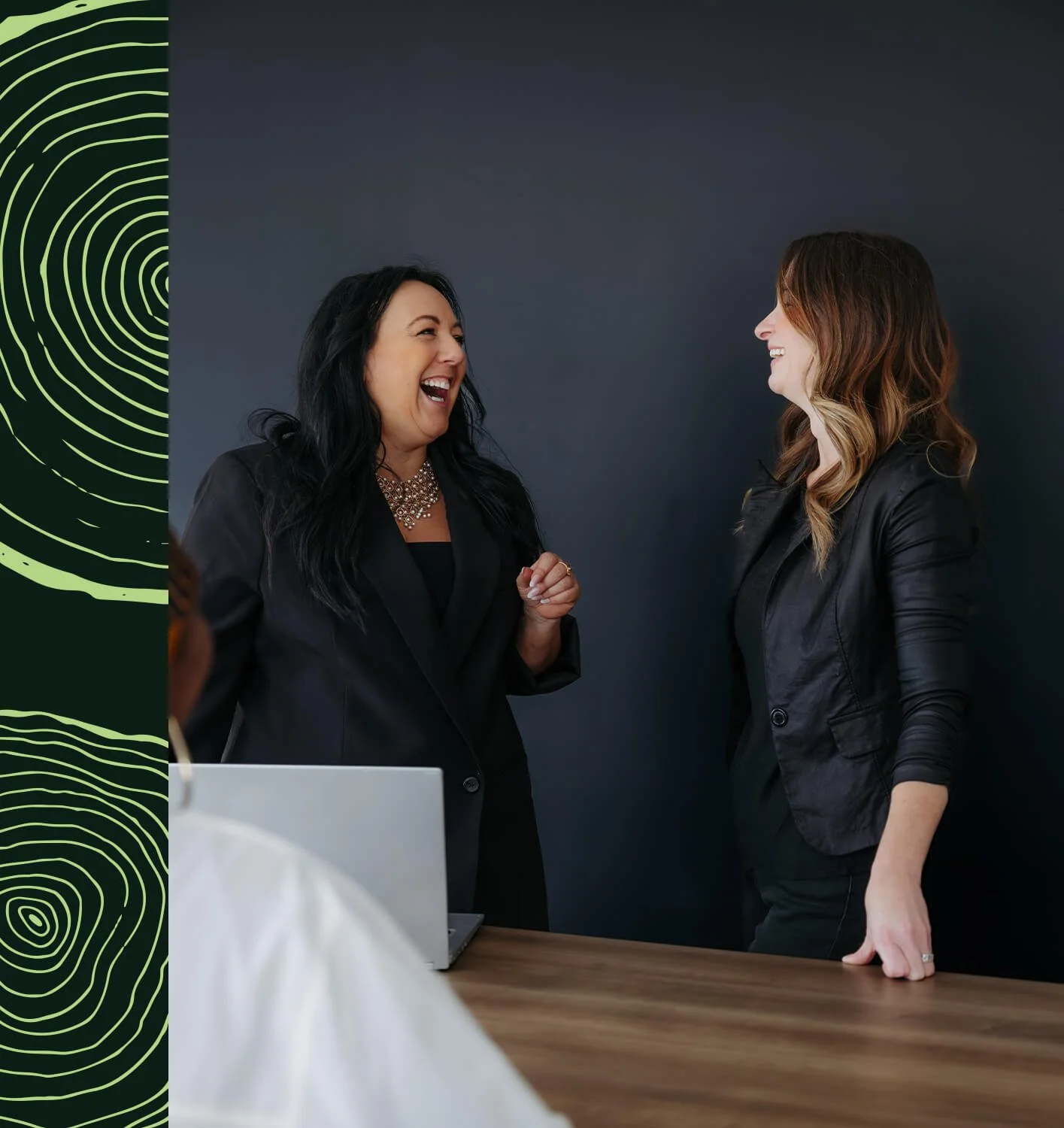 Julie Duhamel et Mélinda Roy souriantes discutent dans un bureau moderne avec un mur noir et un ordinateur portable sur une table en bois.