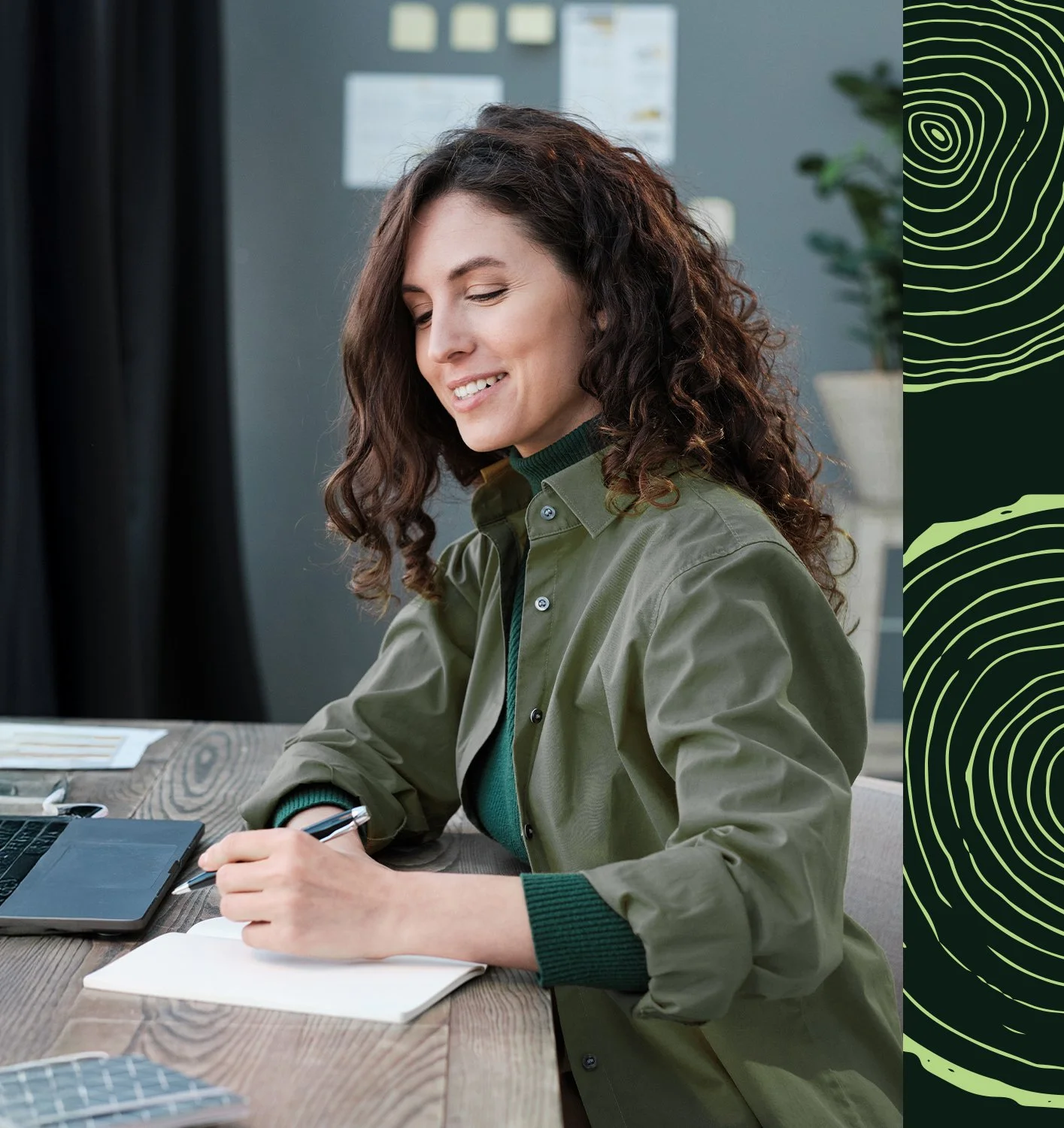 Une femme souriante avec des cheveux bouclés assise à un bureau, en train d'écrire dans un carnet, avec un ordinateur portable et une tablette sur la table.