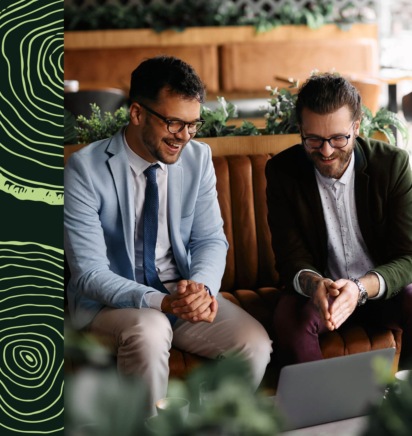 Deux hommes d'affaires habillés formellement, assis sur un sofa en cuir marron, discutant et souriant dans un café ou un espace de coworking, avec un ordinateur portable ouvert devant eux.