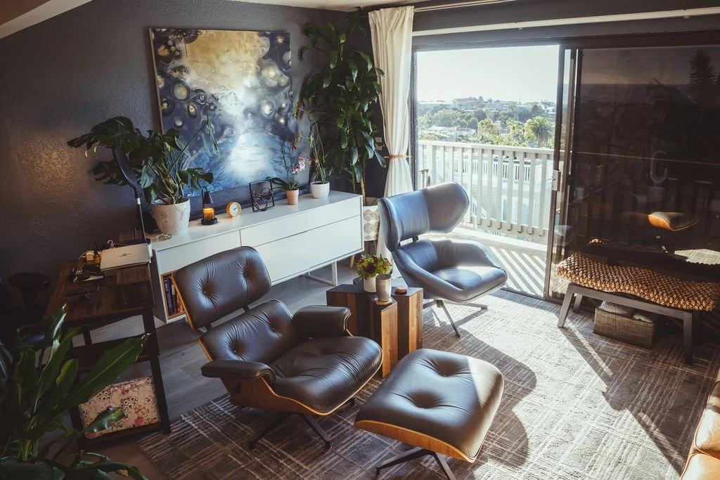 A modern living room with black leather lounge chairs, a white sideboard with plants and decor, a large abstract painting on the wall, and a sliding glass door leading to a balcony with a city view.