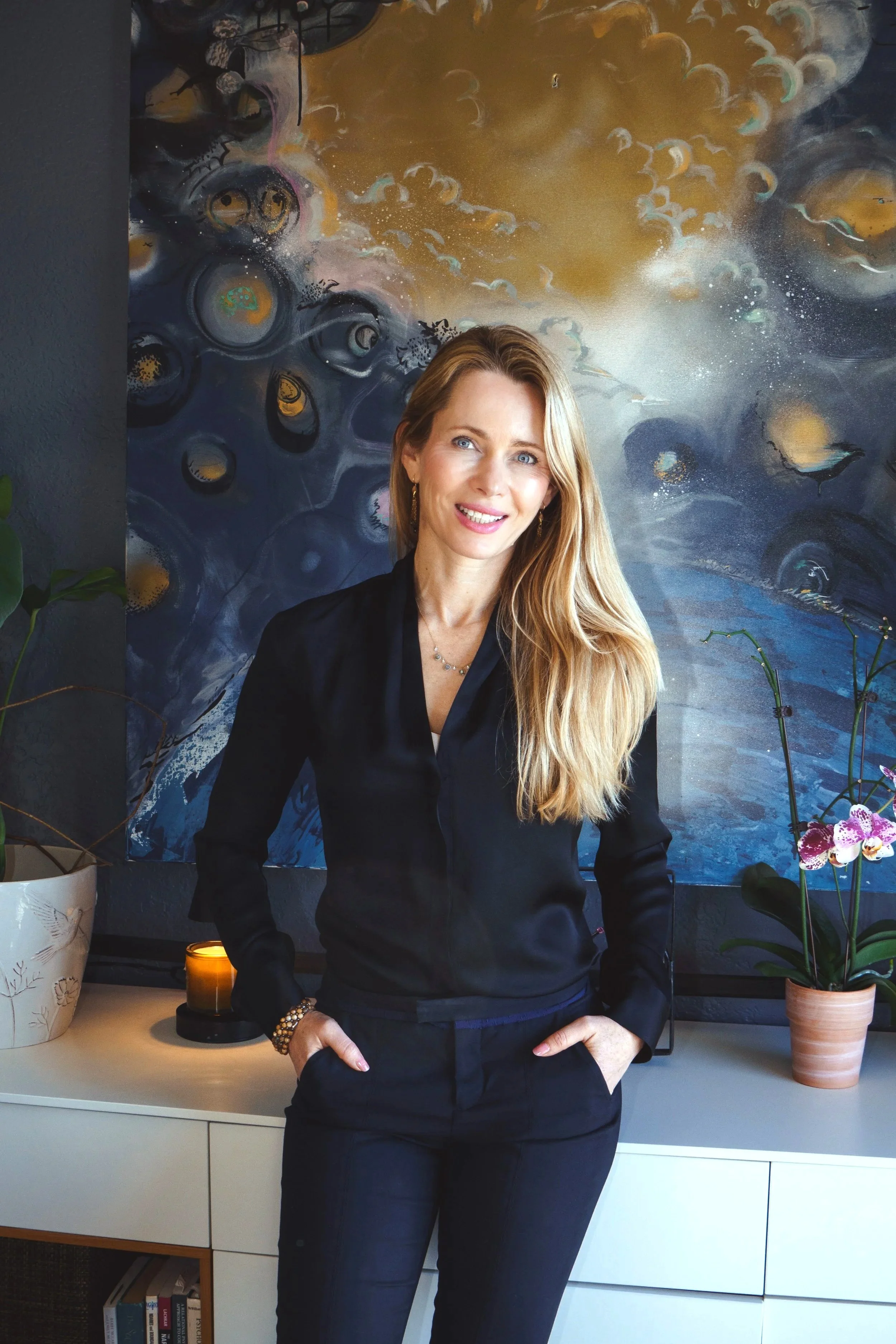 Portrait of a smiling woman with long blonde hair, wearing a black outfit, standing in front of an abstract blue and gold art painting, with nearby potted orchids and a lit candle.
