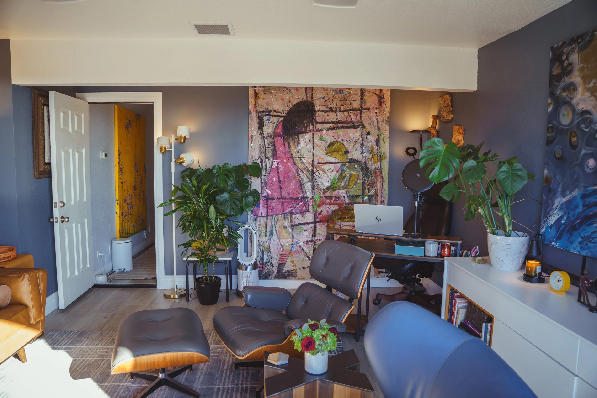 Living room with modern furniture, two large plants, colorful abstract paintings, a desk with a laptop, and various decorative items.