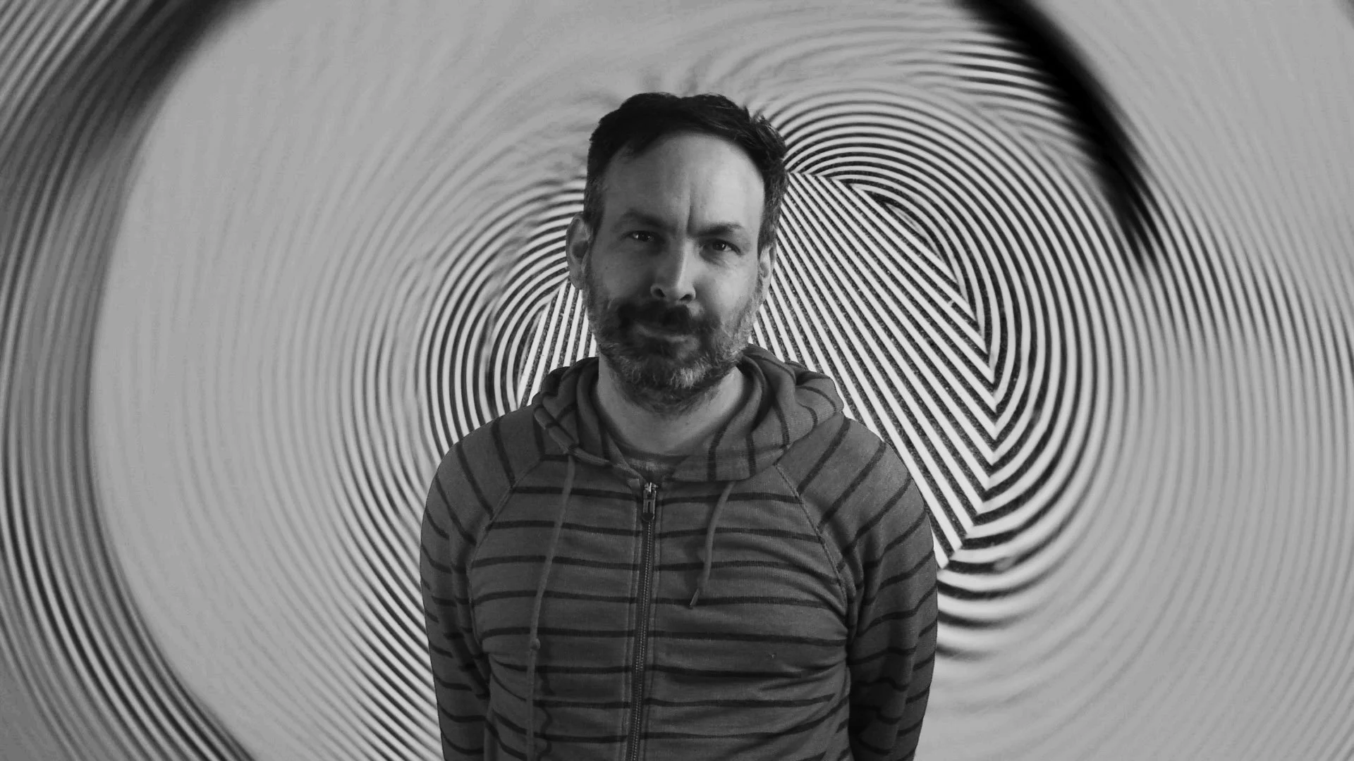 A black and white photograph of a man with a beard smiling and wearing a striped hooded sweatshirt in front of an abstract background with straight and curving lines.