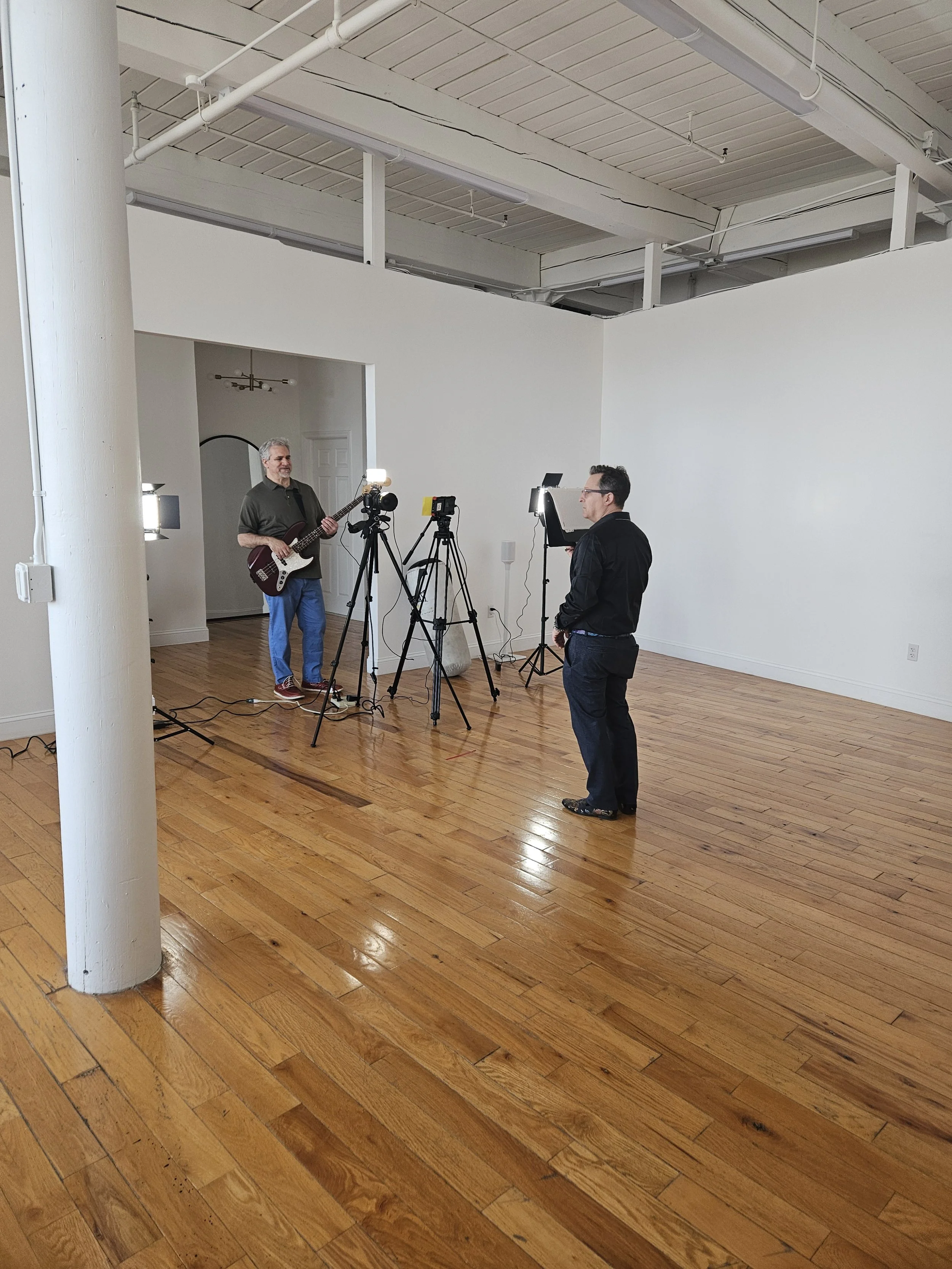 Musician playing guitar while being filmed by a small crew with multiple cameras and lights in a bright studio with hardwood floors and white walls