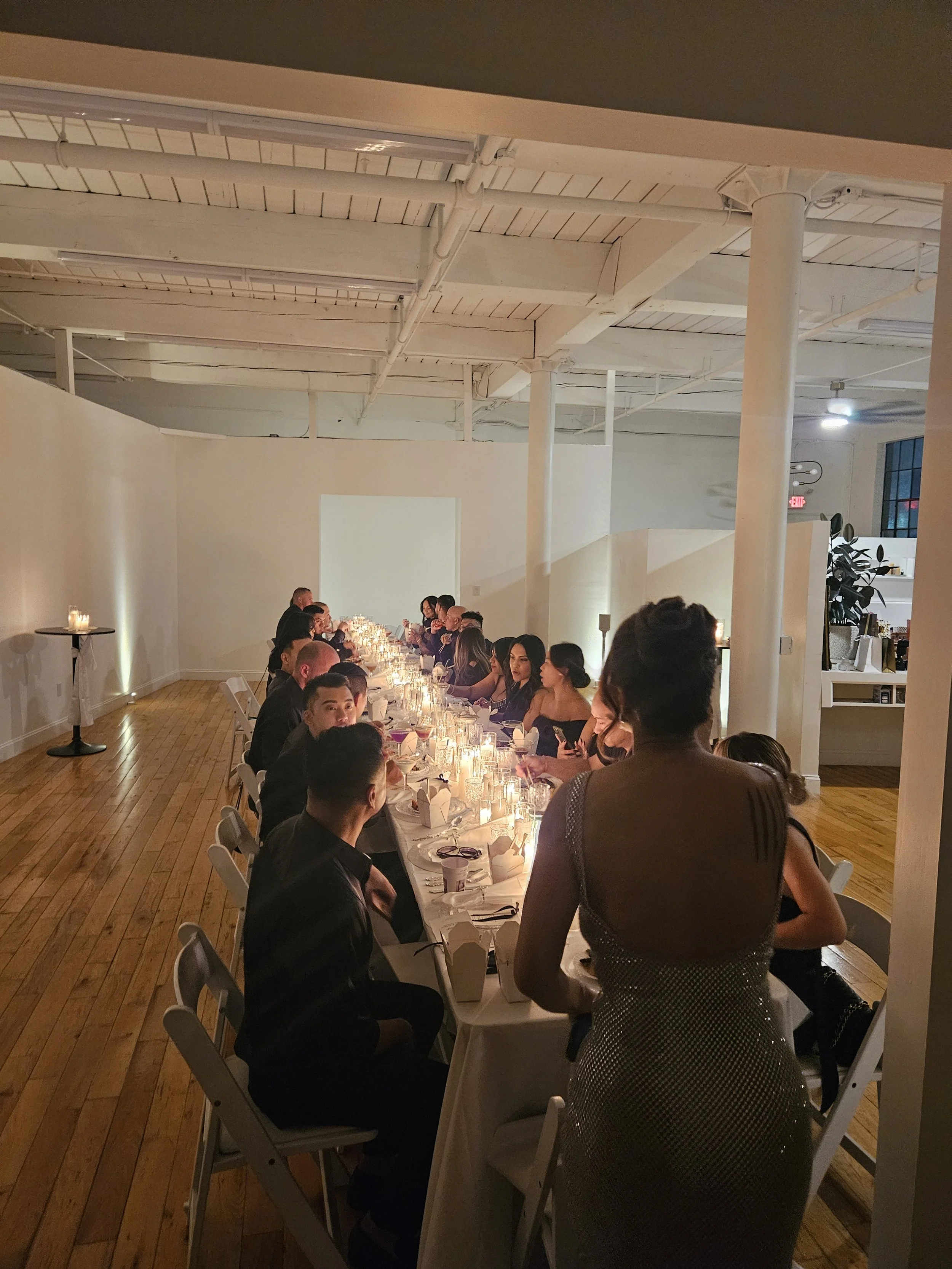 Guests seated at a long candlelit dinner table in a loft-style venue with white walls, columns, and warm lighting